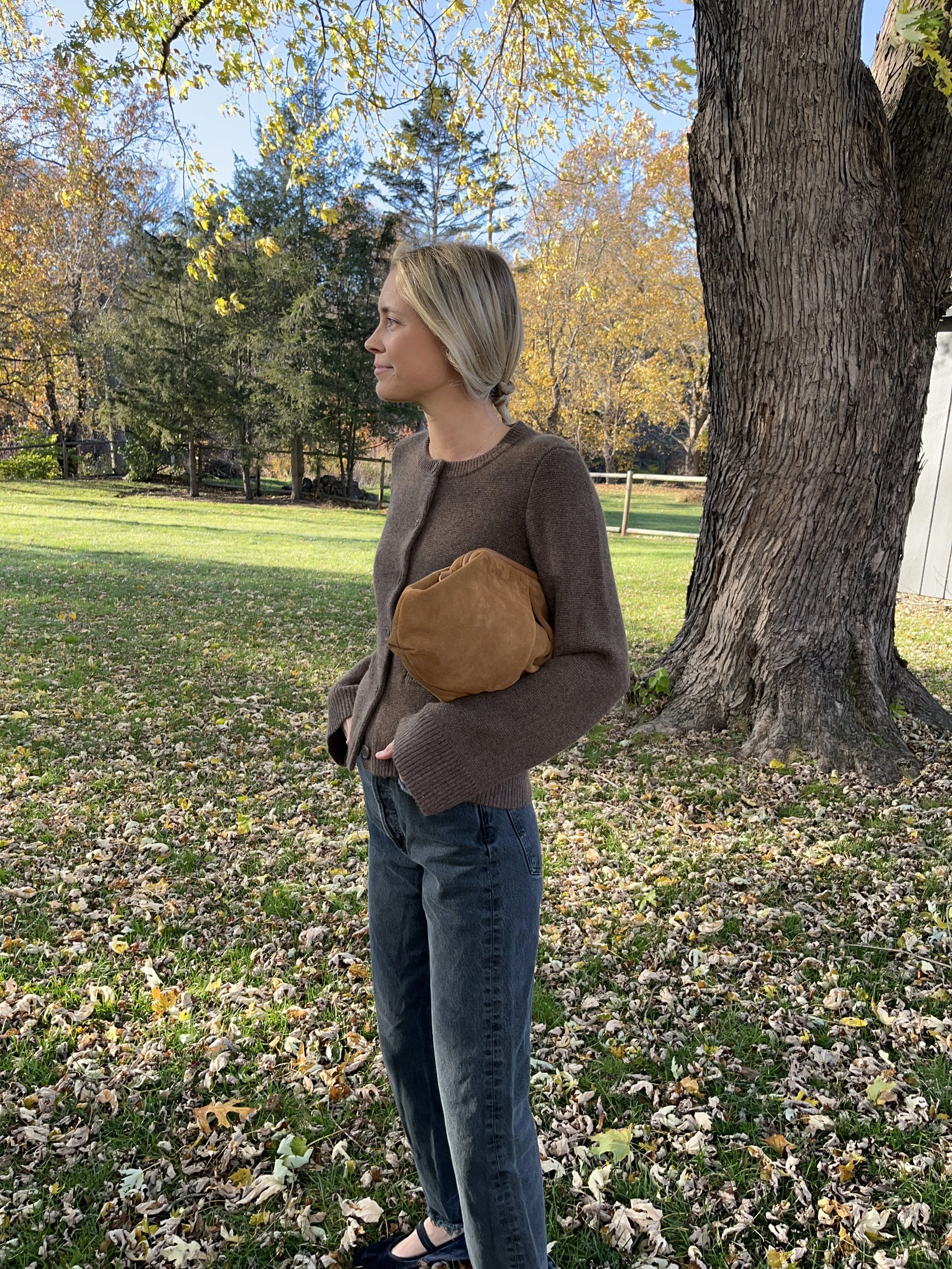 A woman standing outdoors in a park during autumn, holding a brown clutch bag, wearing a brown cardigan, blue jeans, and black shoes, with trees and fallen leaves in the background.