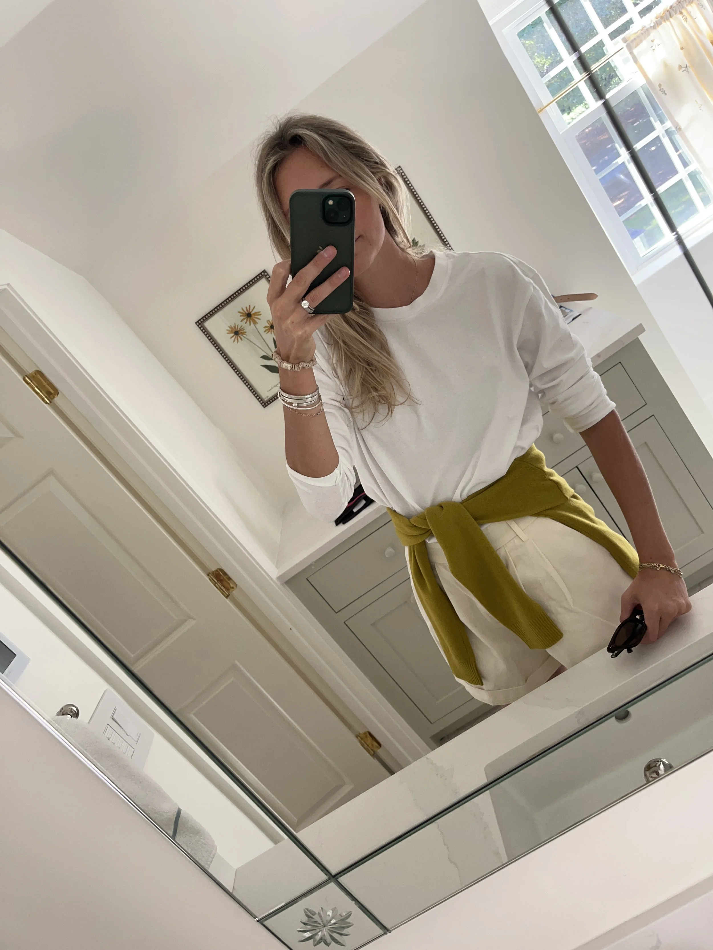A woman taking a mirror selfie in a spacious, well-lit living room with hardwood floors, large windows, a white sofa, and a side table with a plant.