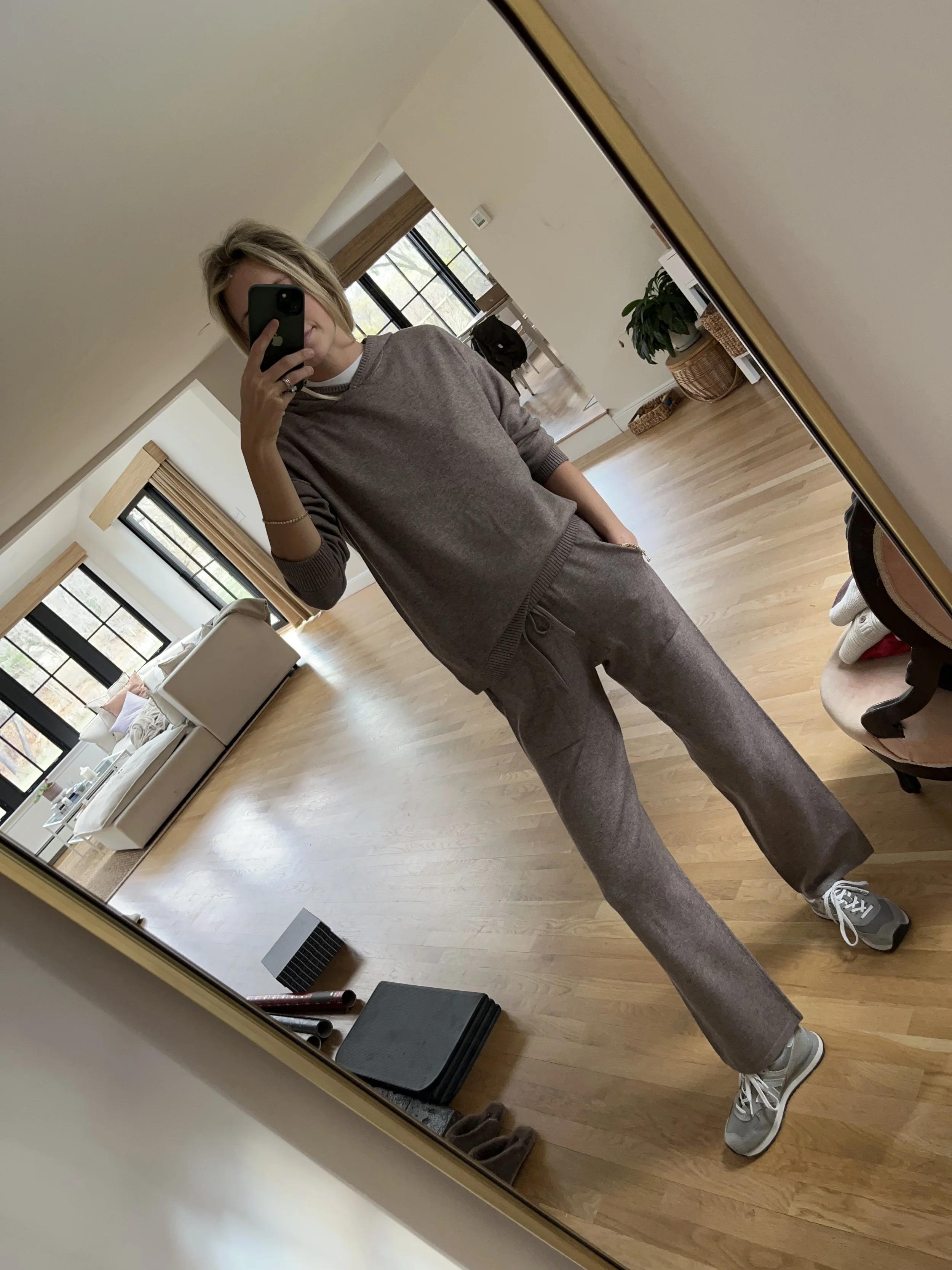 Person taking a mirror selfie in a living room with hardwood floors, wearing a black fleece jacket, gray shirt, black leggings, gray sneakers, and a white cap with a logo.