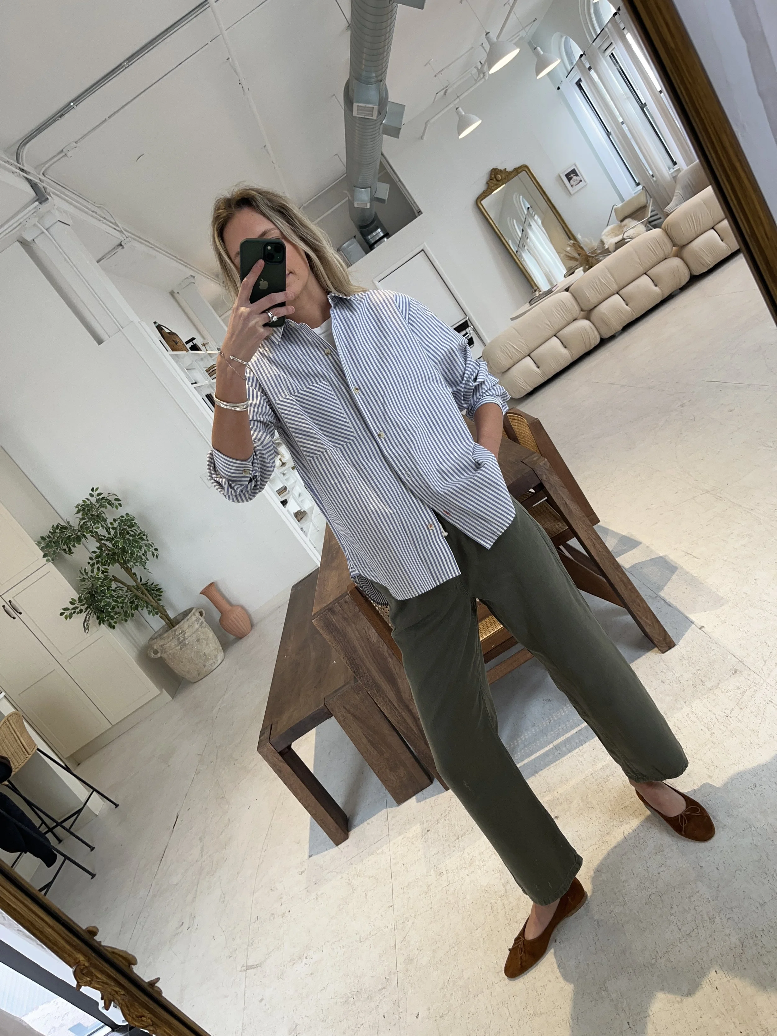 A woman taking a mirror selfie in a spacious living room with wooden floors, large windows, and minimal furniture, wearing a beige jacket, black top, blue jeans, and tan shoes.