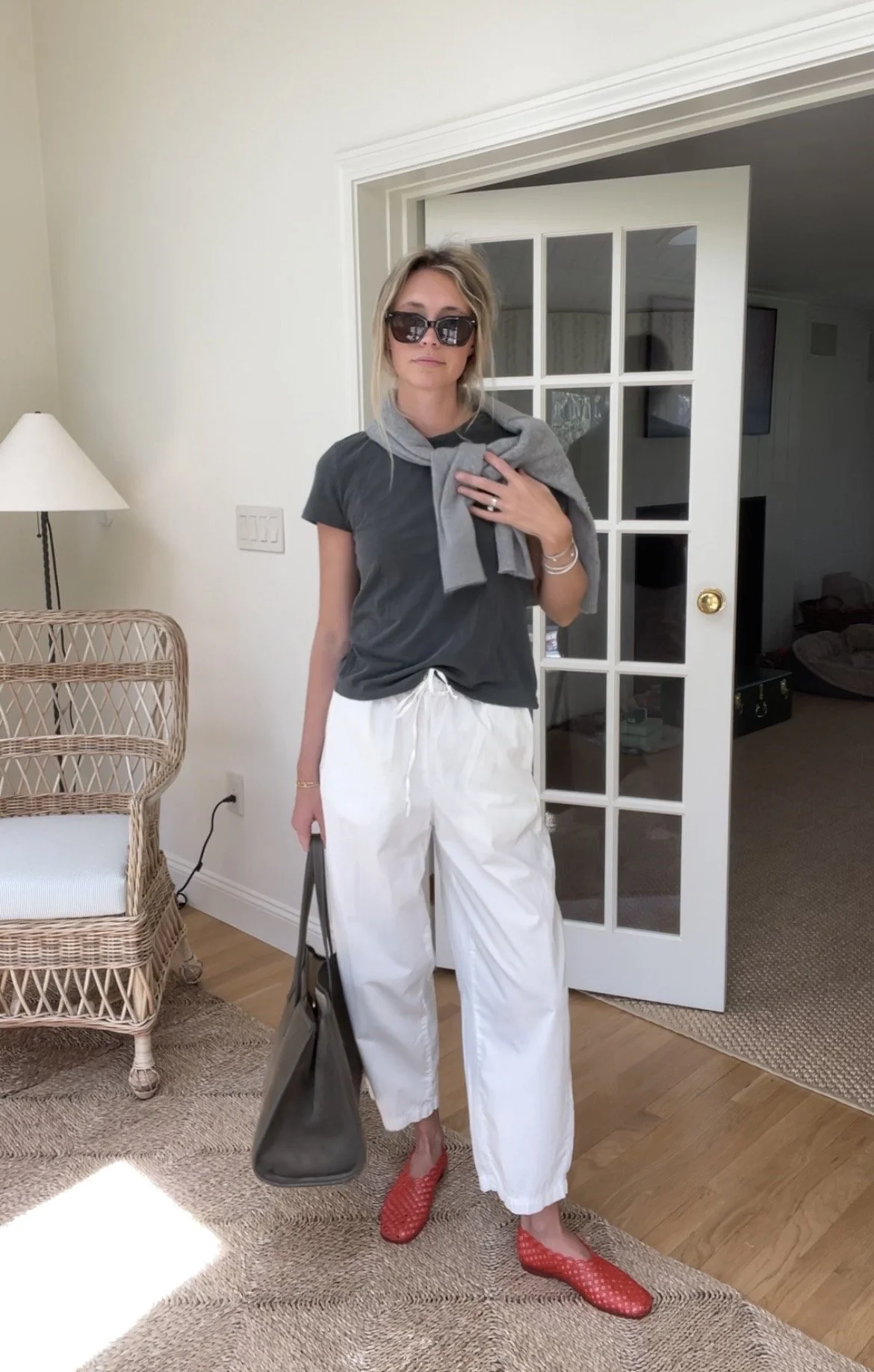 Person taking a mirror selfie in a living room with hardwood floors, wearing a black fleece jacket, gray shirt, black leggings, gray sneakers, and a white cap with a logo.