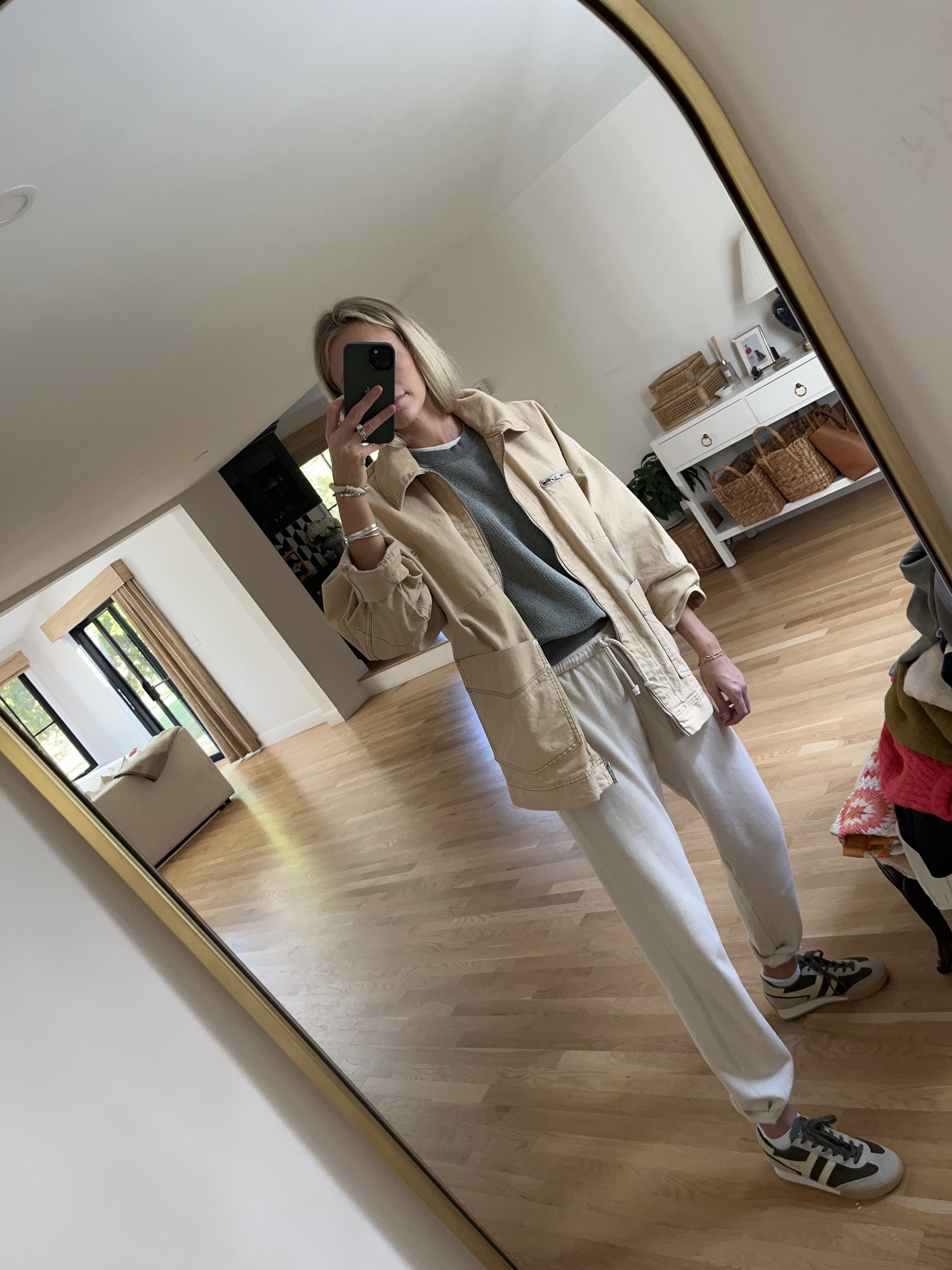 Person taking a mirror selfie in a spacious, well-lit living room with wooden floors, white furniture, and large windows.