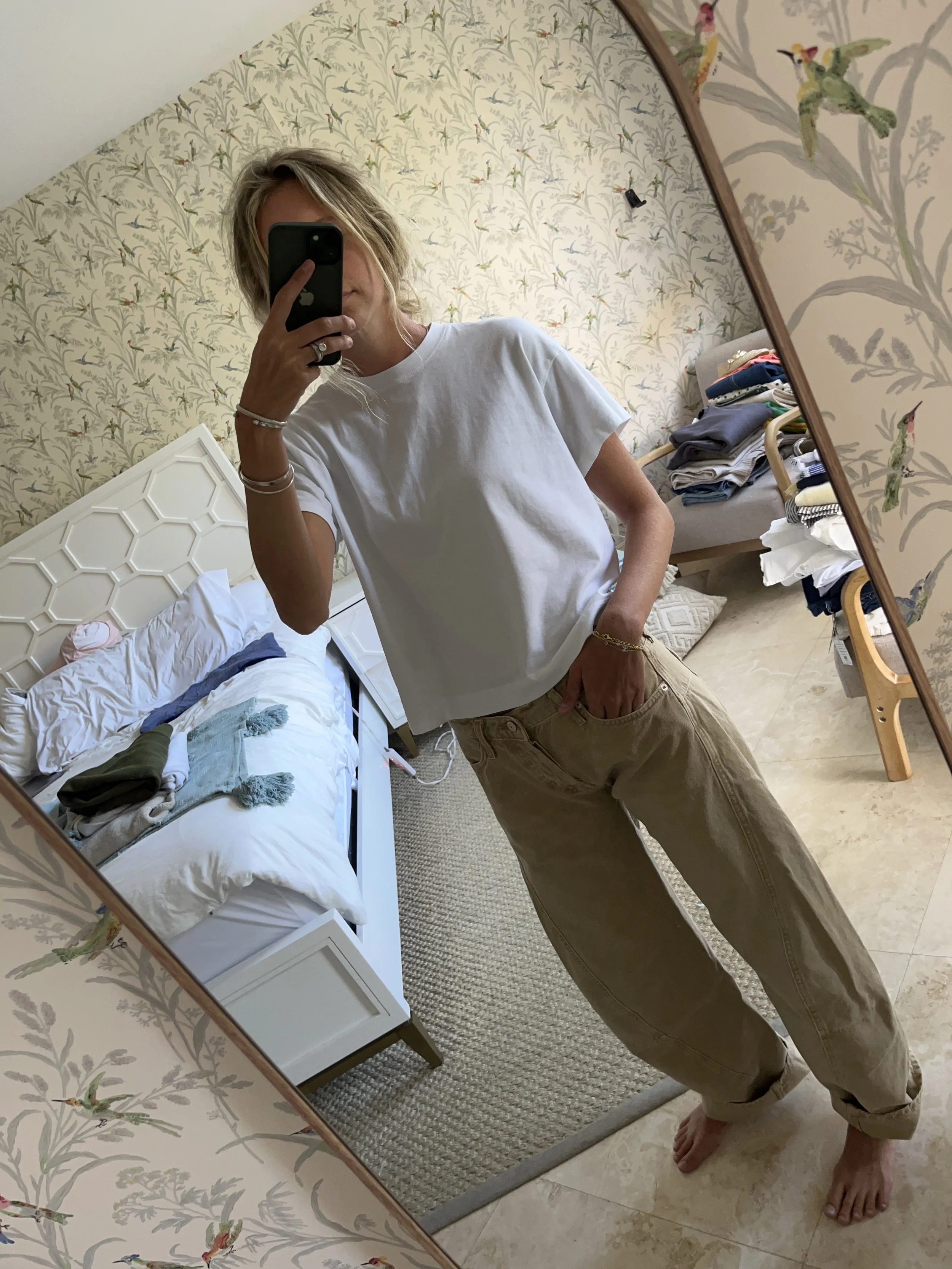 A woman taking a mirror selfie in a spacious, well-lit living room with hardwood floors, large windows, a white sofa, and a side table with a plant.