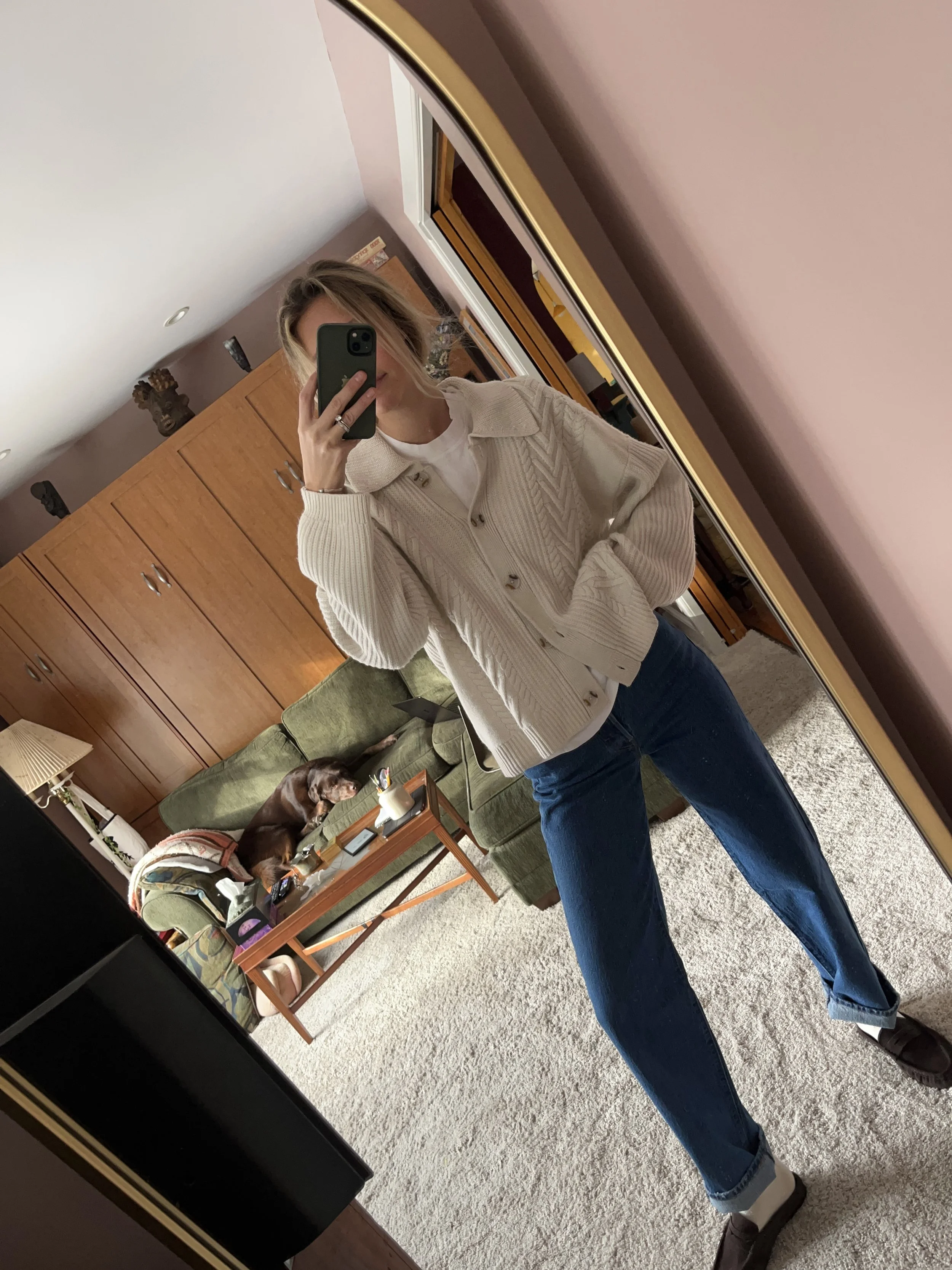 Person taking a mirror selfie in a room with wooden floors, wearing a striped brown and white long-sleeve shirt, light blue jeans, and black and white sneakers.