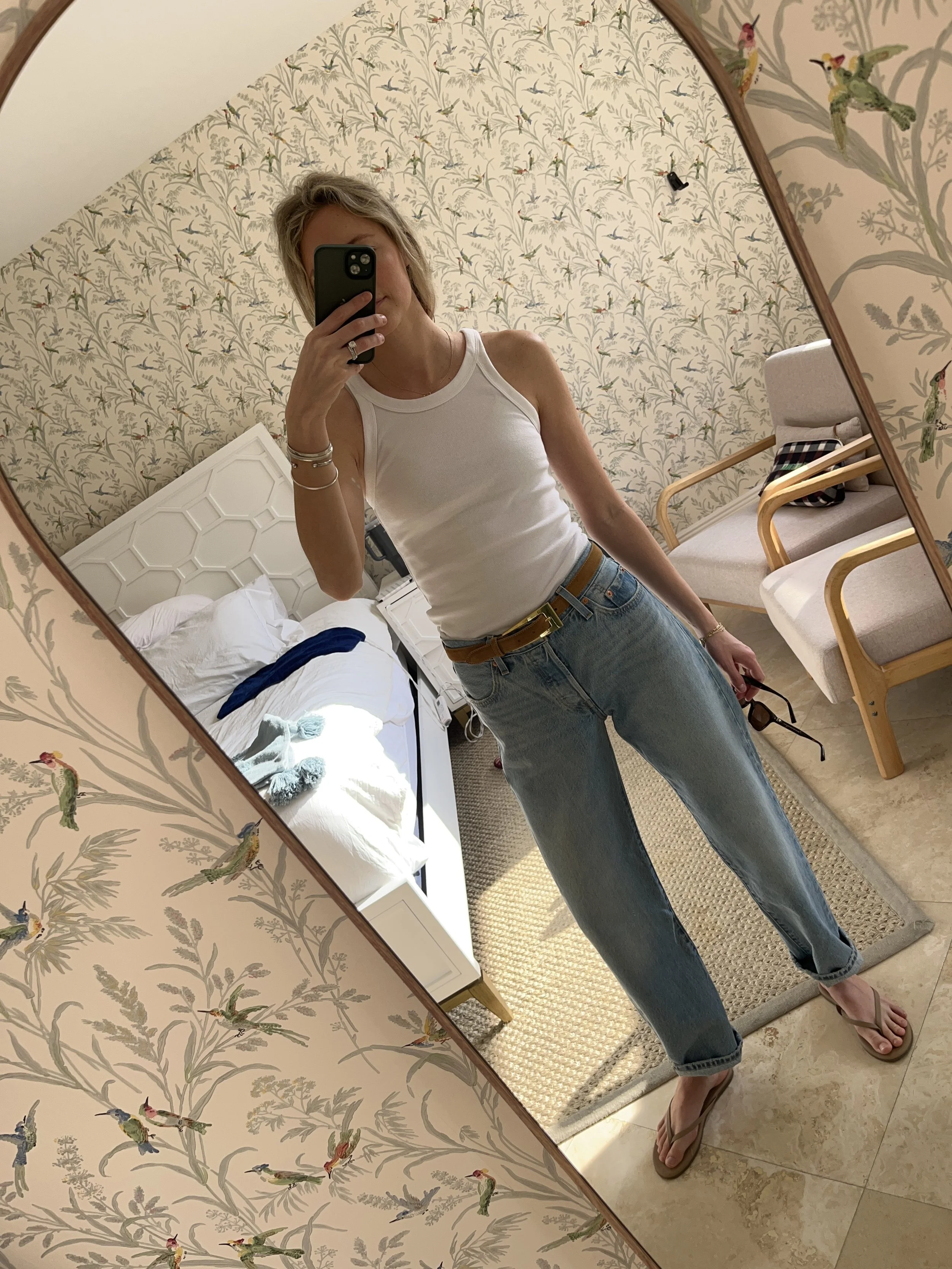 Person taking a mirror selfie in a room with wooden floors, wearing a striped brown and white long-sleeve shirt, light blue jeans, and black and white sneakers.