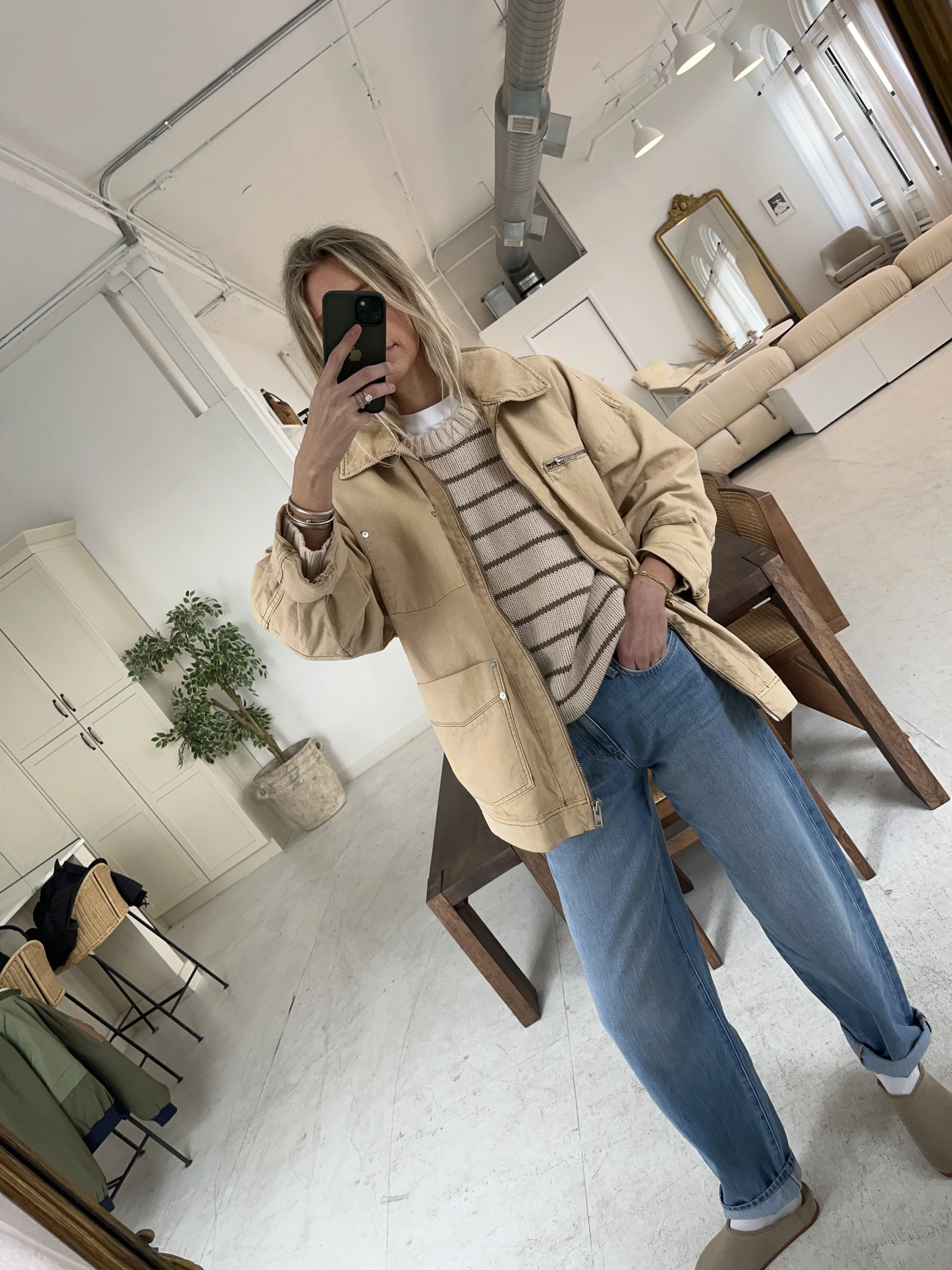 Person taking a mirror selfie in a room with wooden floors, wearing a striped brown and white long-sleeve shirt, light blue jeans, and black and white sneakers.
