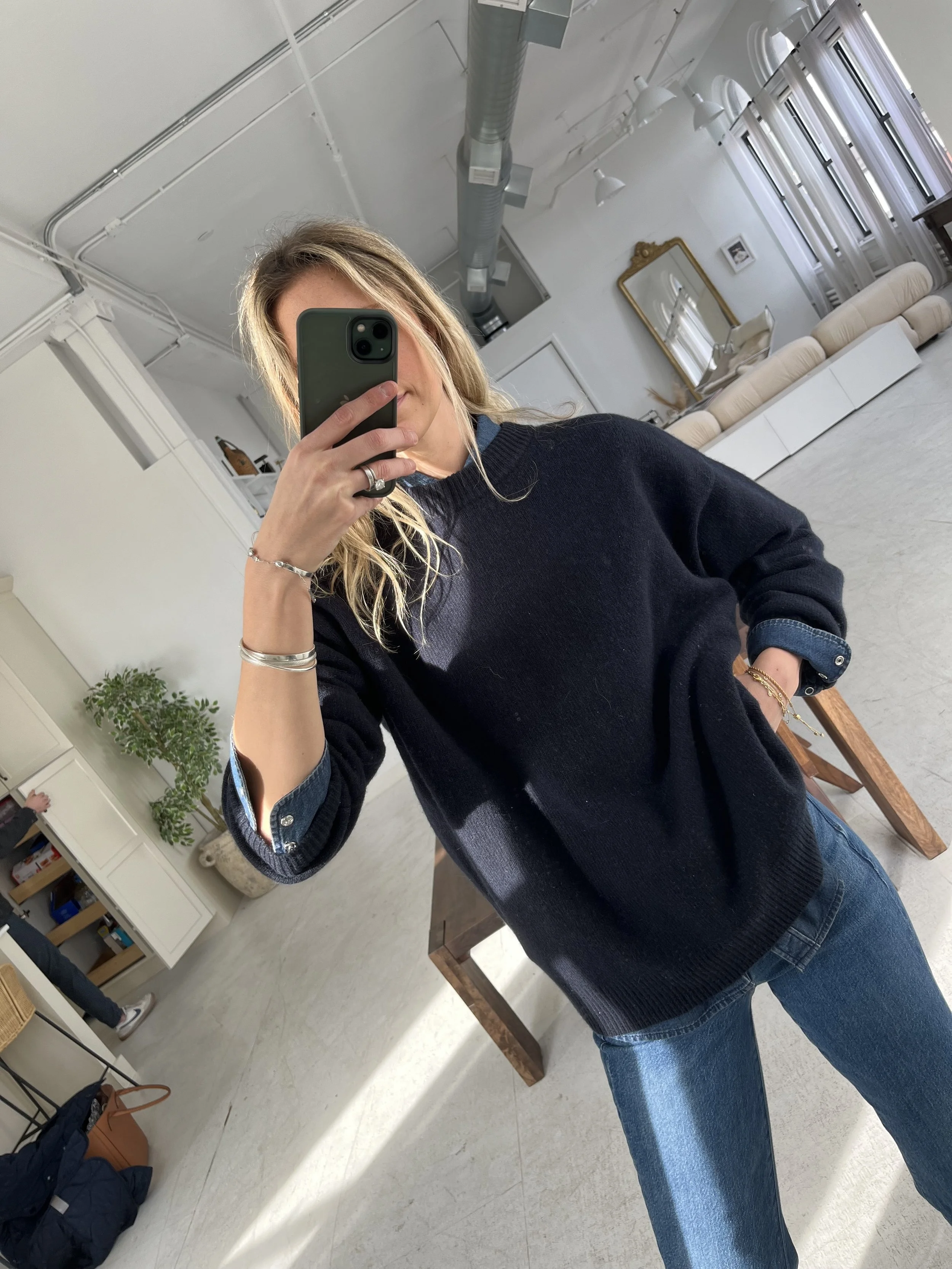 A woman taking a mirror selfie in a spacious, well-lit living room with hardwood floors, large windows, a white sofa, and a side table with a plant.