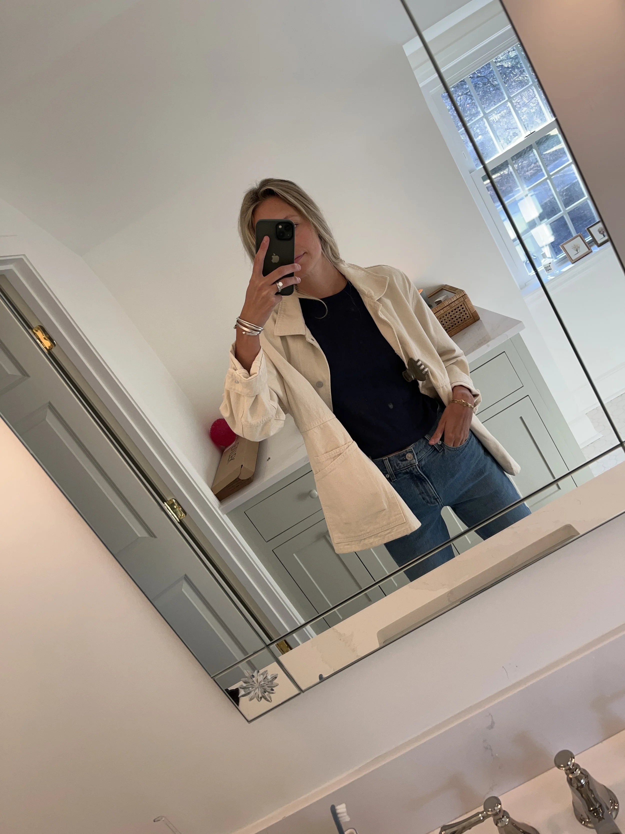 A woman taking a mirror selfie in a spacious, well-lit living room with hardwood floors, large windows, a white sofa, and a side table with a plant.