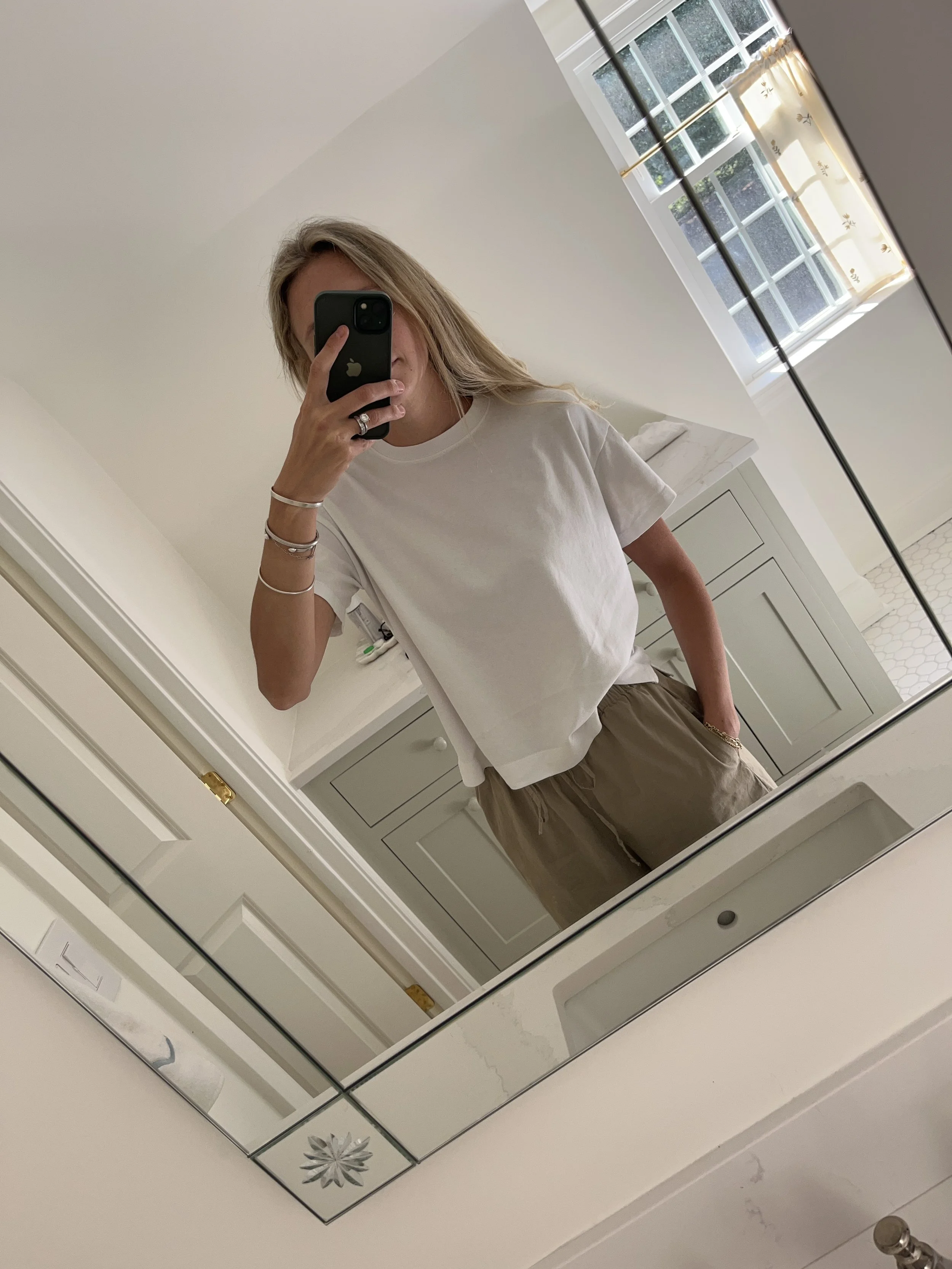 A woman taking a mirror selfie in a spacious, well-lit living room with hardwood floors, large windows, a white sofa, and a side table with a plant.