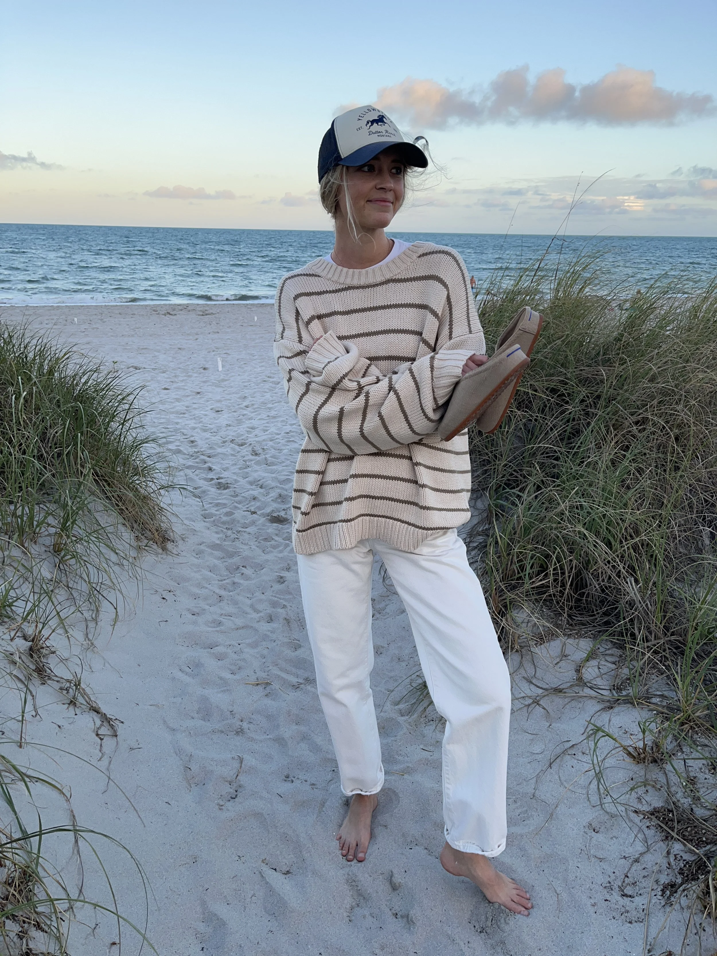 Person taking a mirror selfie in a living room with hardwood floors, wearing a black fleece jacket, gray shirt, black leggings, gray sneakers, and a white cap with a logo.