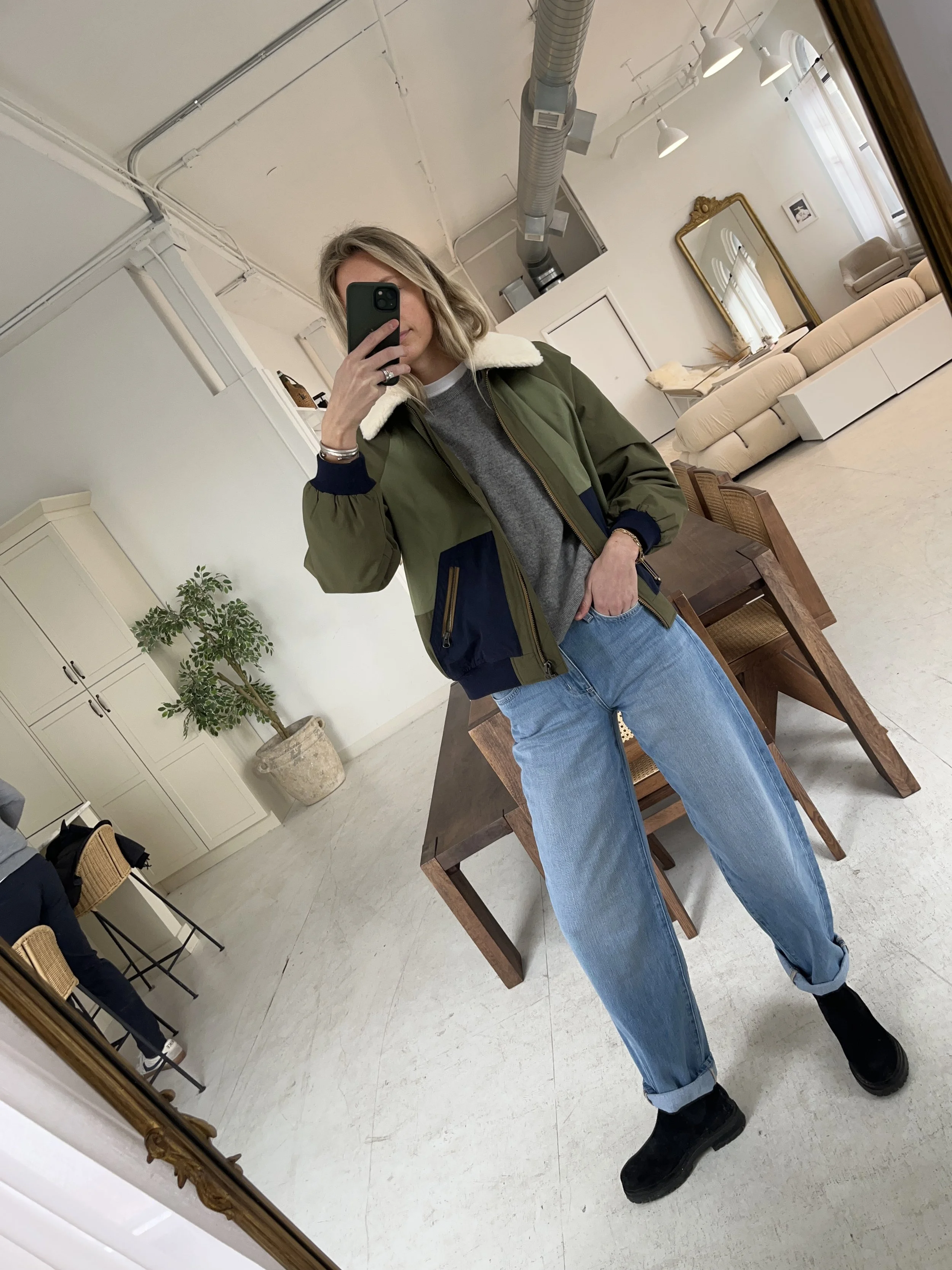 Person taking a mirror selfie in a room with wooden floors, wearing a striped brown and white long-sleeve shirt, light blue jeans, and black and white sneakers.