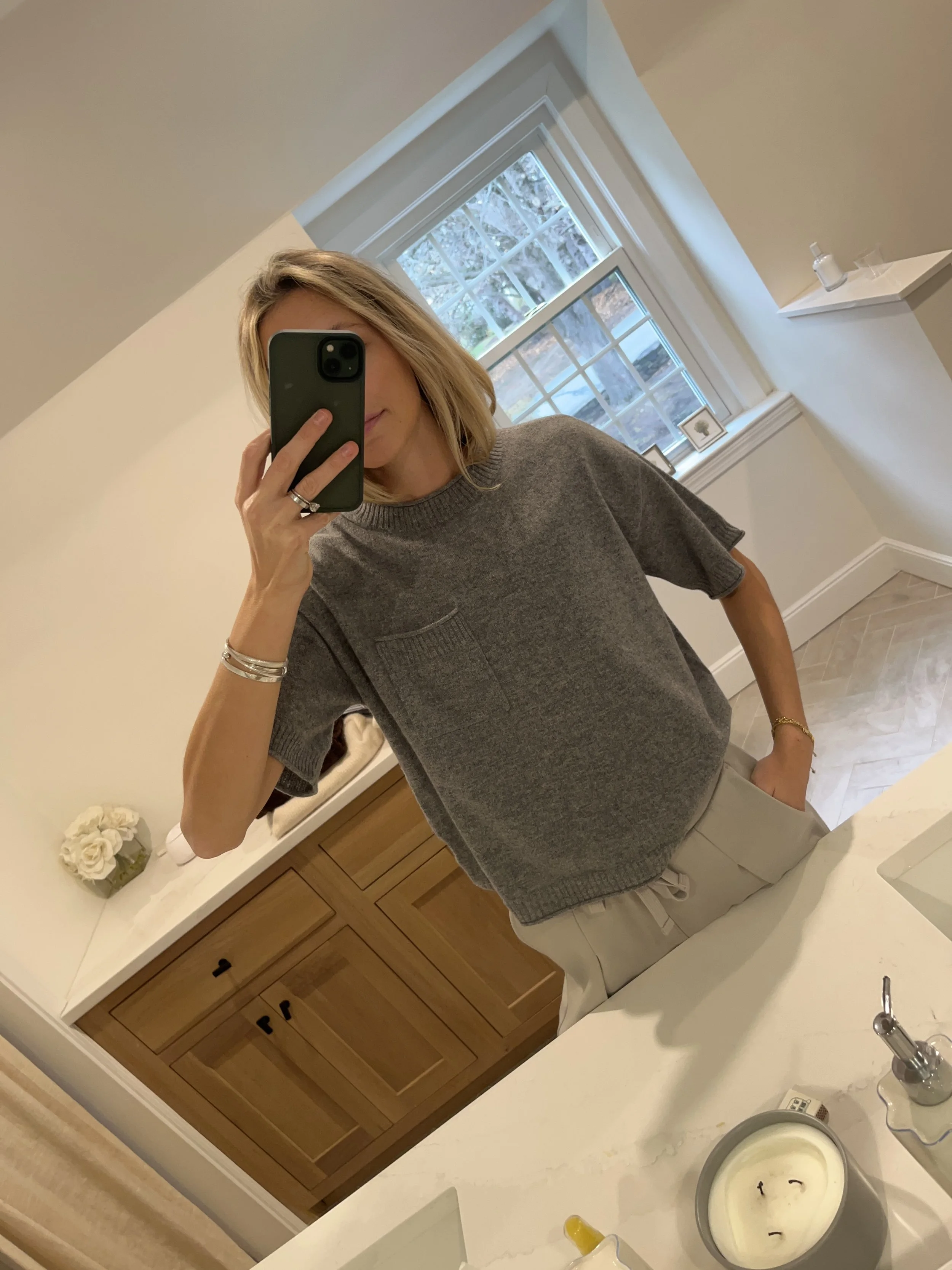 Person taking a mirror selfie in a room with wooden floors, wearing a striped brown and white long-sleeve shirt, light blue jeans, and black and white sneakers.