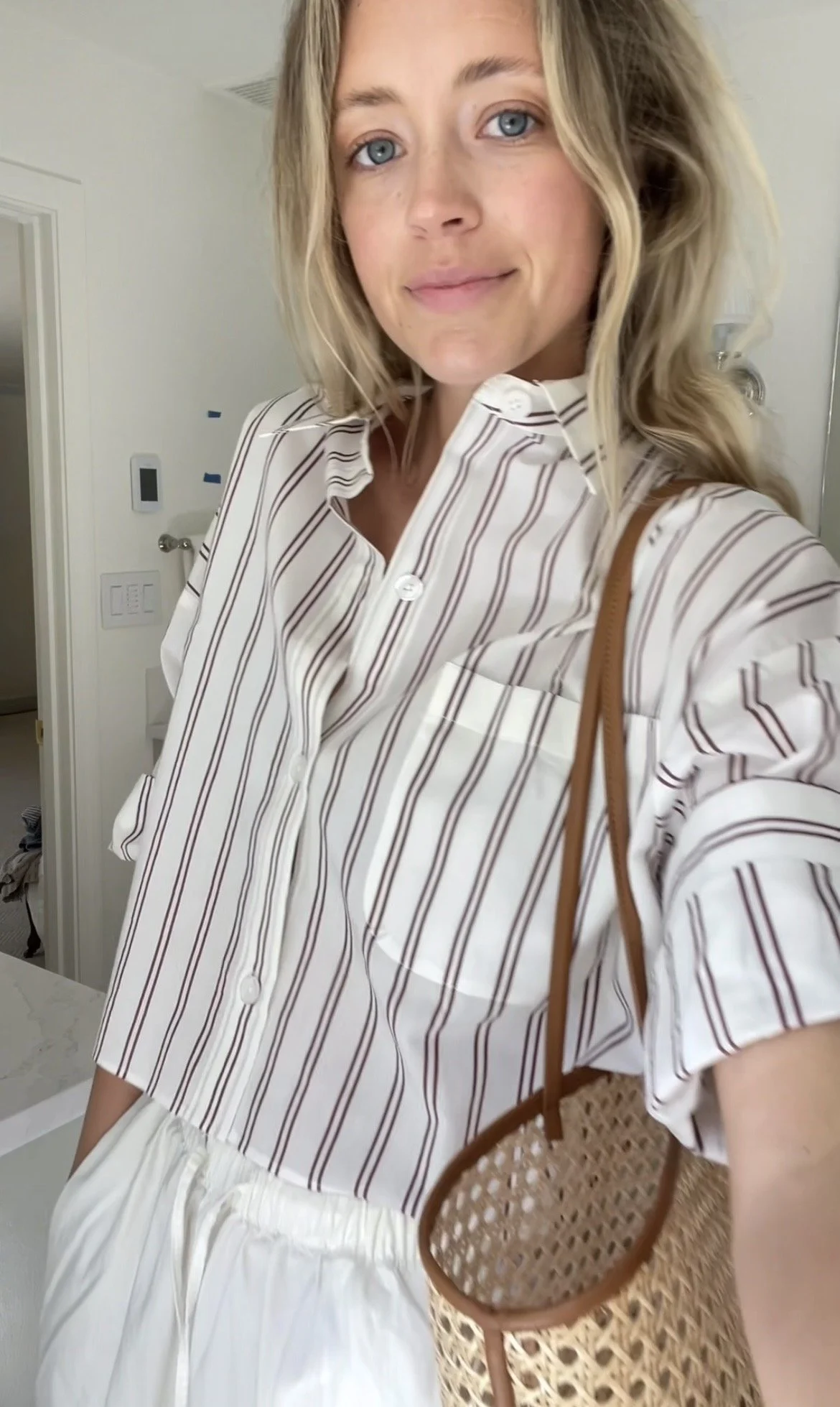 A woman taking a mirror selfie in a spacious living room with wooden floors, large windows, and minimal furniture, wearing a beige jacket, black top, blue jeans, and tan shoes.
