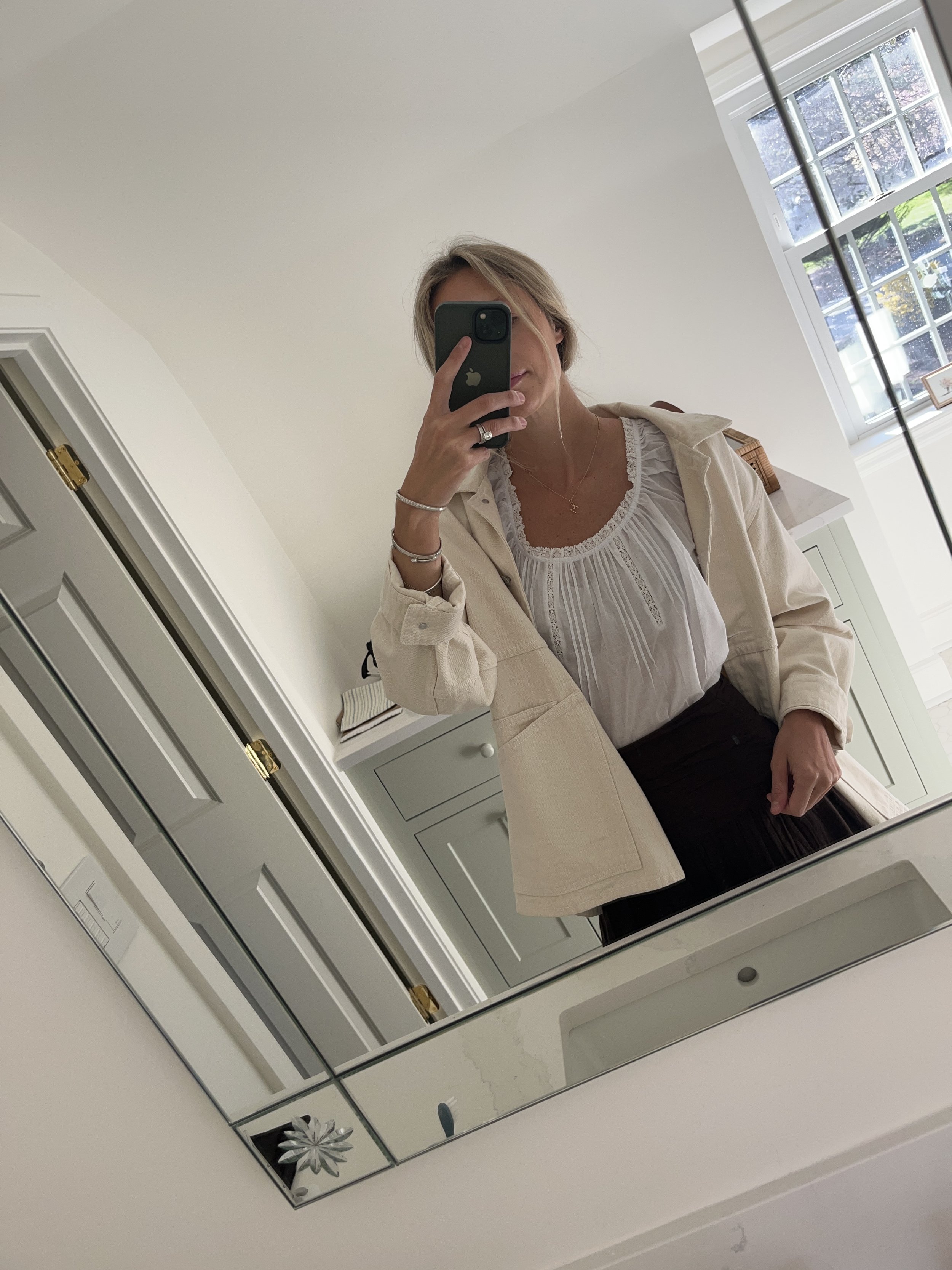Person taking a mirror selfie in a living room with hardwood floors, wearing a black fleece jacket, gray shirt, black leggings, gray sneakers, and a white cap with a logo.