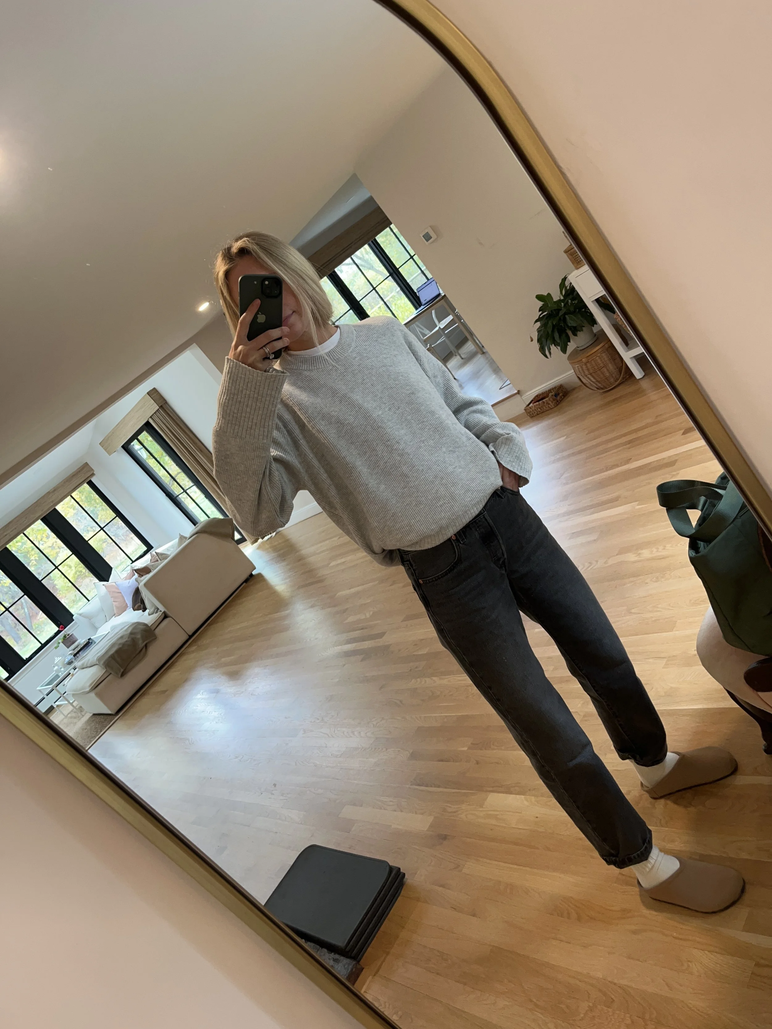 A person taking a mirror selfie in a living room with wooden floors, a white sofa, large black-framed windows, and houseplants, wearing a light gray sweater, black jeans, and beige slippers.