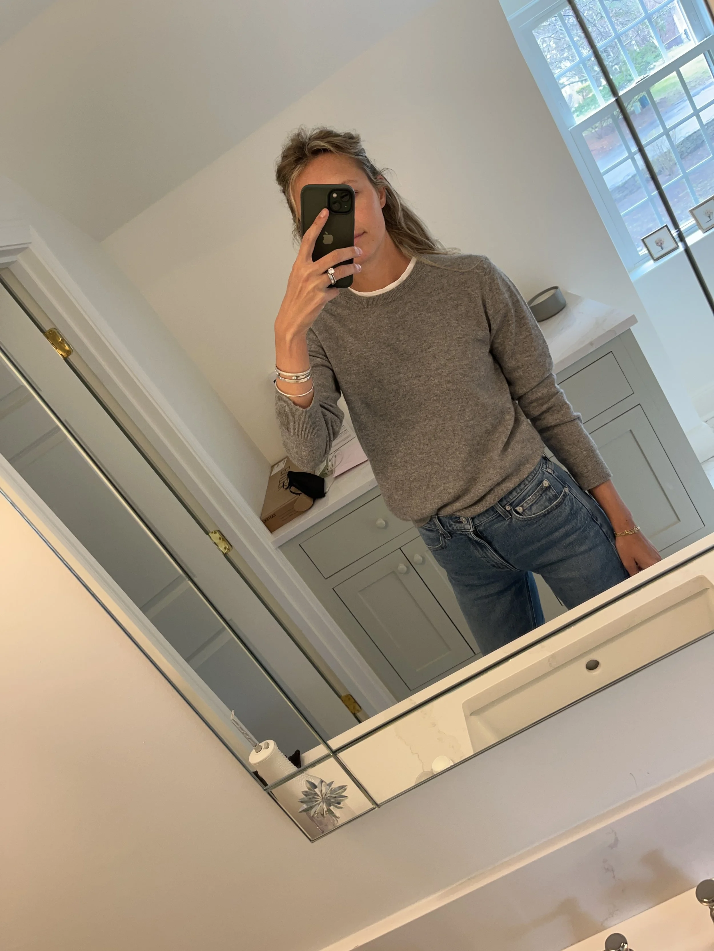 Person taking a mirror selfie in a room with wooden floors, wearing a striped brown and white long-sleeve shirt, light blue jeans, and black and white sneakers.