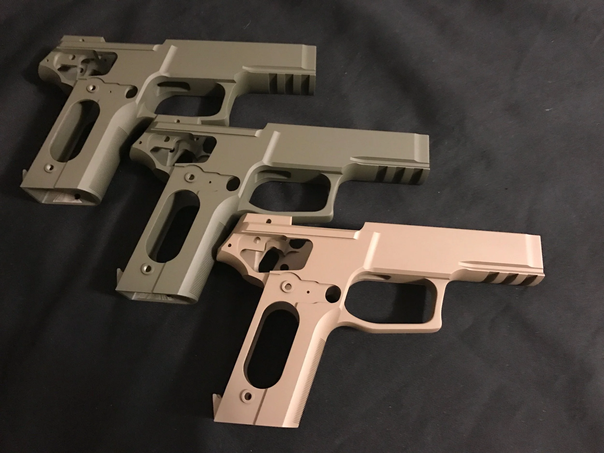 Three handgun frames in different colors, two in a tan shade and one in a lighter pinkish shade, laid on a dark surface.