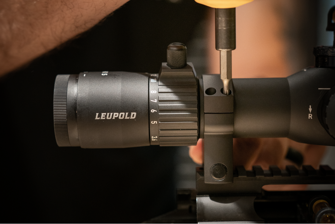 Close-up of a rifle scope marked 'Leupold,' mounted on a firearm, with a screwdriver being used to adjust the screw near the scope.