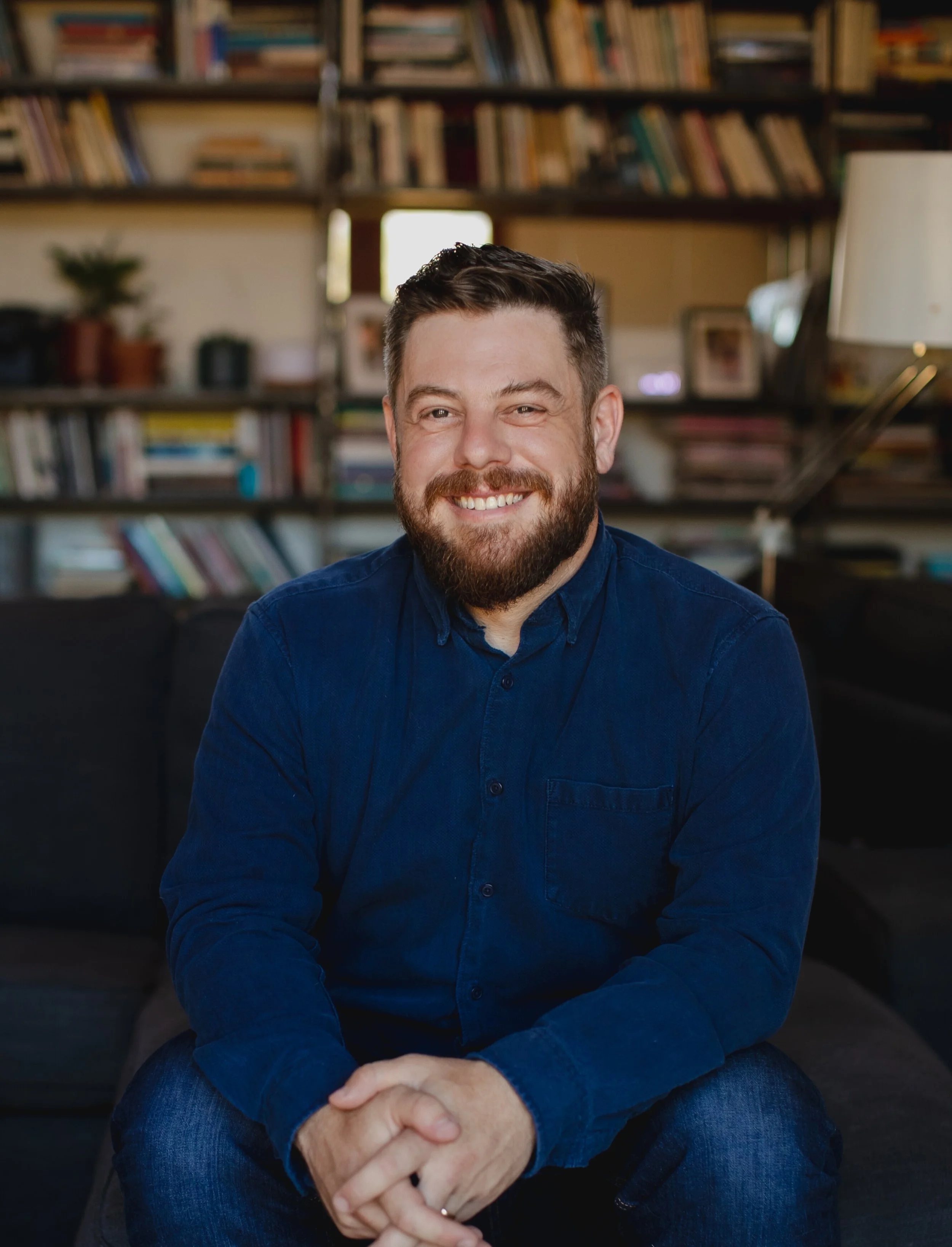 A man with a beard and short dark hair sitting indoors in front of a bookshelf filled with books. He is smiling and wearing a navy blue button-up shirt and blue jeans.