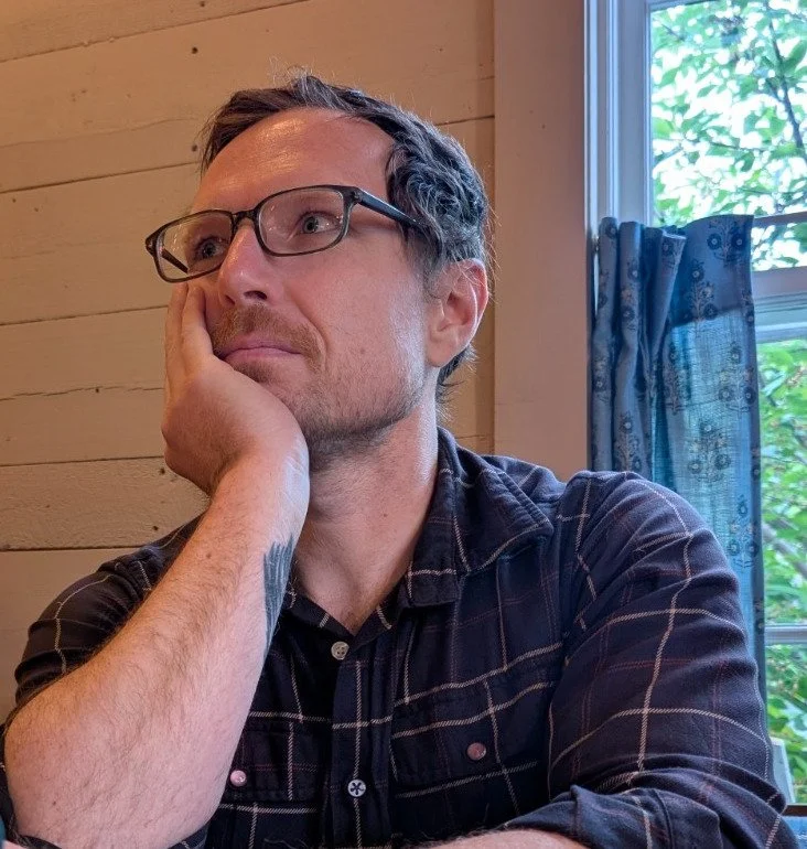 A man with glasses and short dark hair is sitting indoors, resting his head on his hand, looking thoughtful. He is wearing a dark plaid shirt, and there is a window with a blue curtain behind him, showing trees outside.