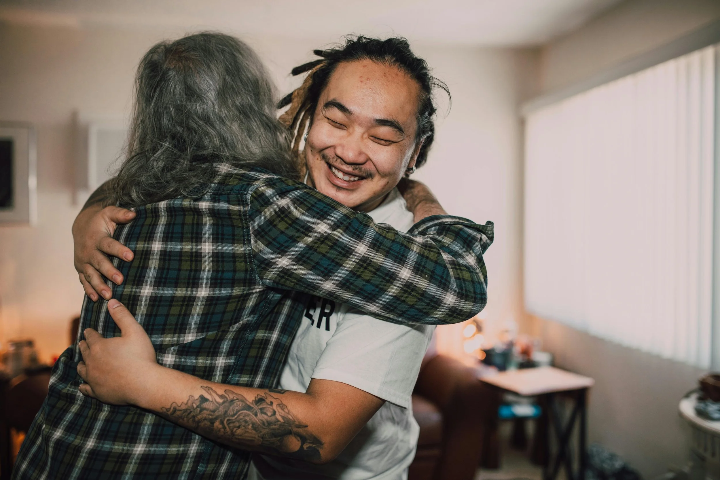 Two people embracing warmly in a cozy room, one wearing a plaid shirt and the other with visible tattoos on their arm in an act of kindness.