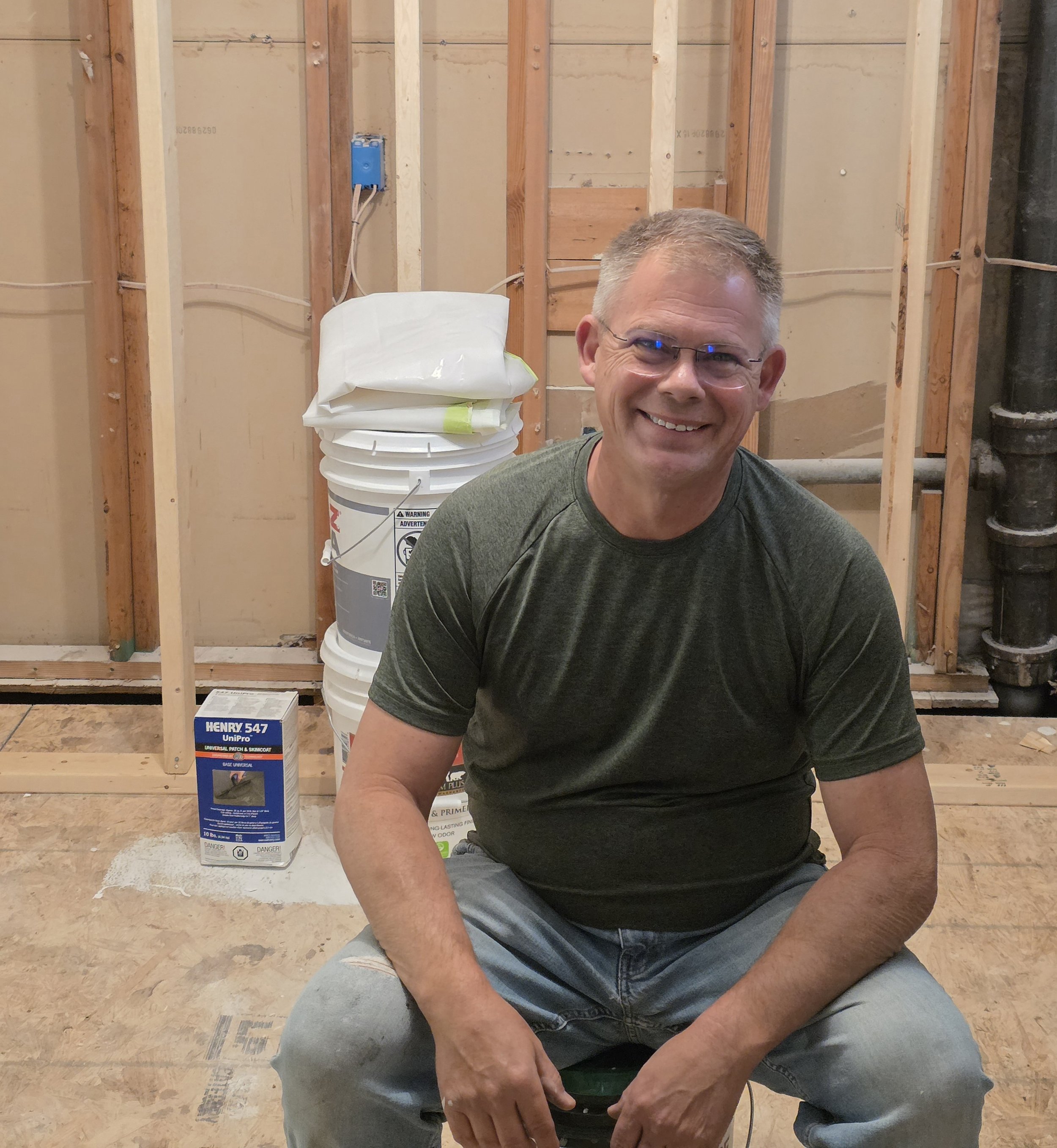 A smiling man wearing glasses and a green shirt, crouching in a construction site with exposed wooden studs and pipes in the background.