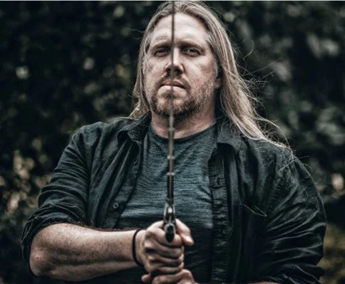 A man with long hair and a beard, holding a sword pointed at the camera, standing outdoors with a blurred forest background.