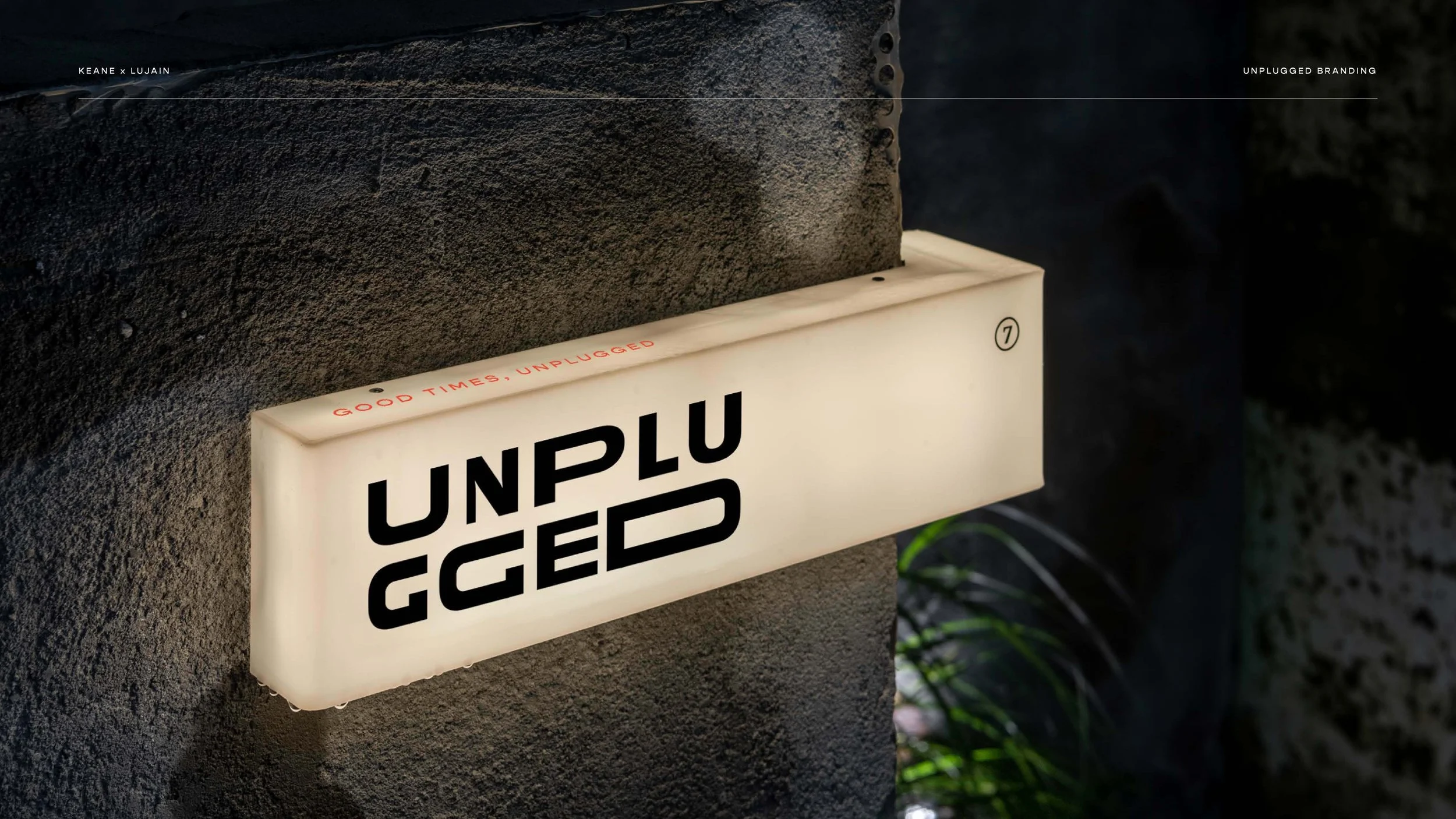 Illuminated sign with the word "UNPLUGGED" in bold black letters on a white background, mounted on a dark textured wall with greenery visible in the background.