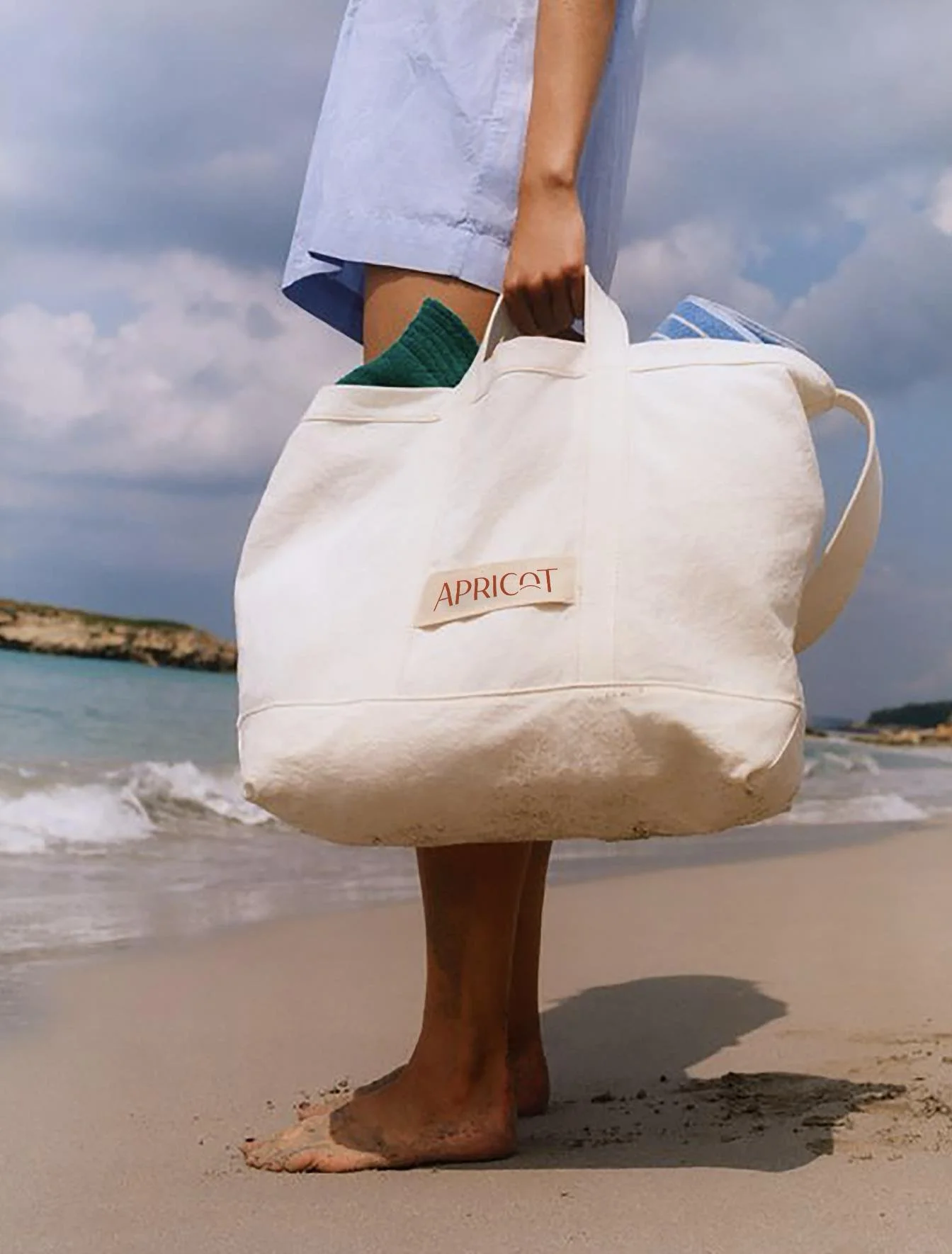 Close-up of a person standing barefoot on a sandy beach, holding a large white tote bag labeled "Apricot" with a blue towel inside, under a cloudy sky.