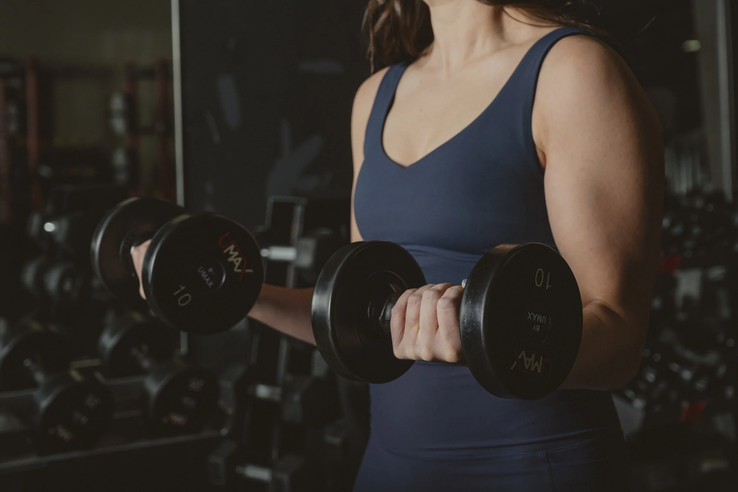 A woman in a blue tank top lifting black dumbbells in a gym with weight racks in the background.