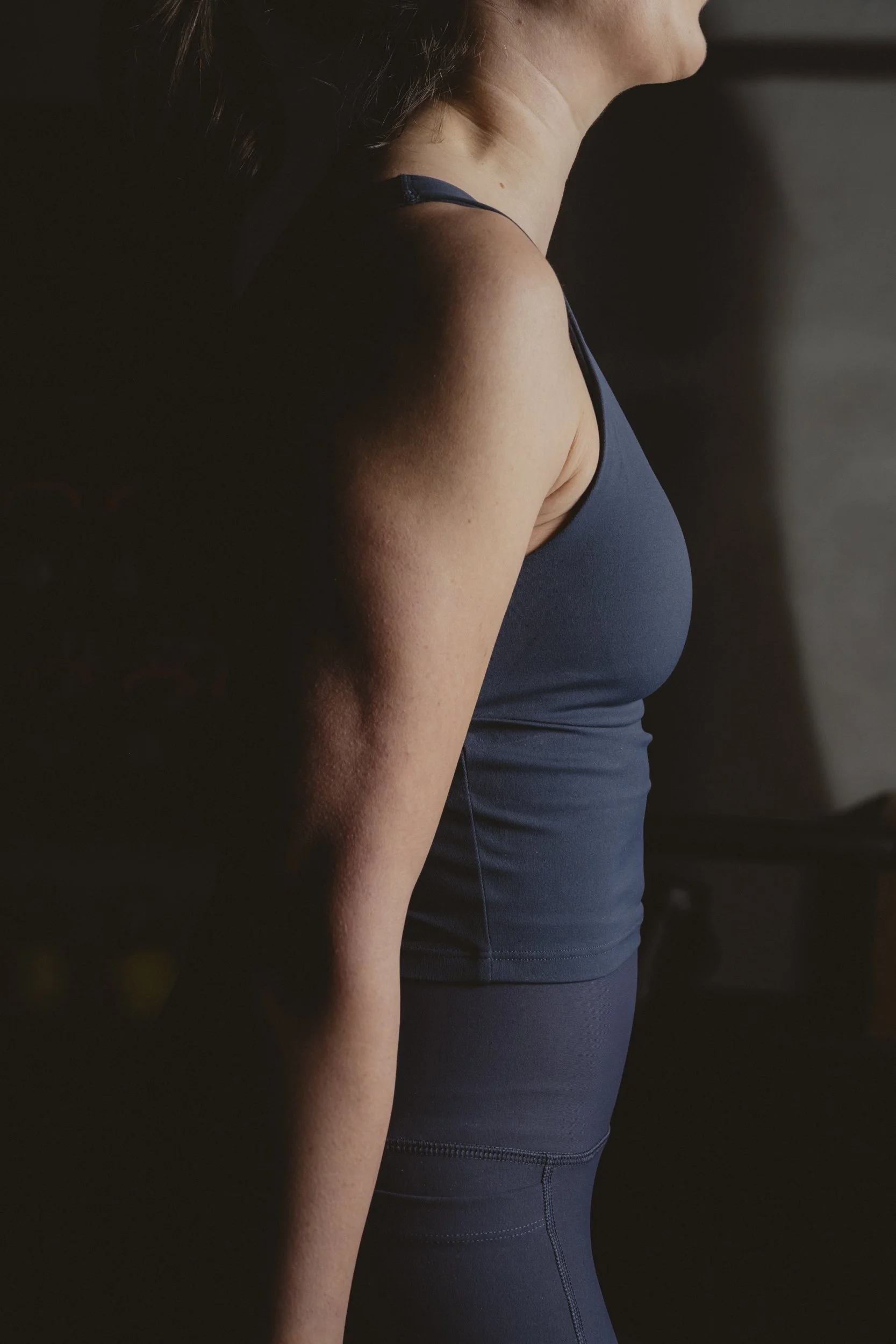 A woman in workout clothes standing in a dimly lit room, with sunlight casting shadows on her arm and torso.