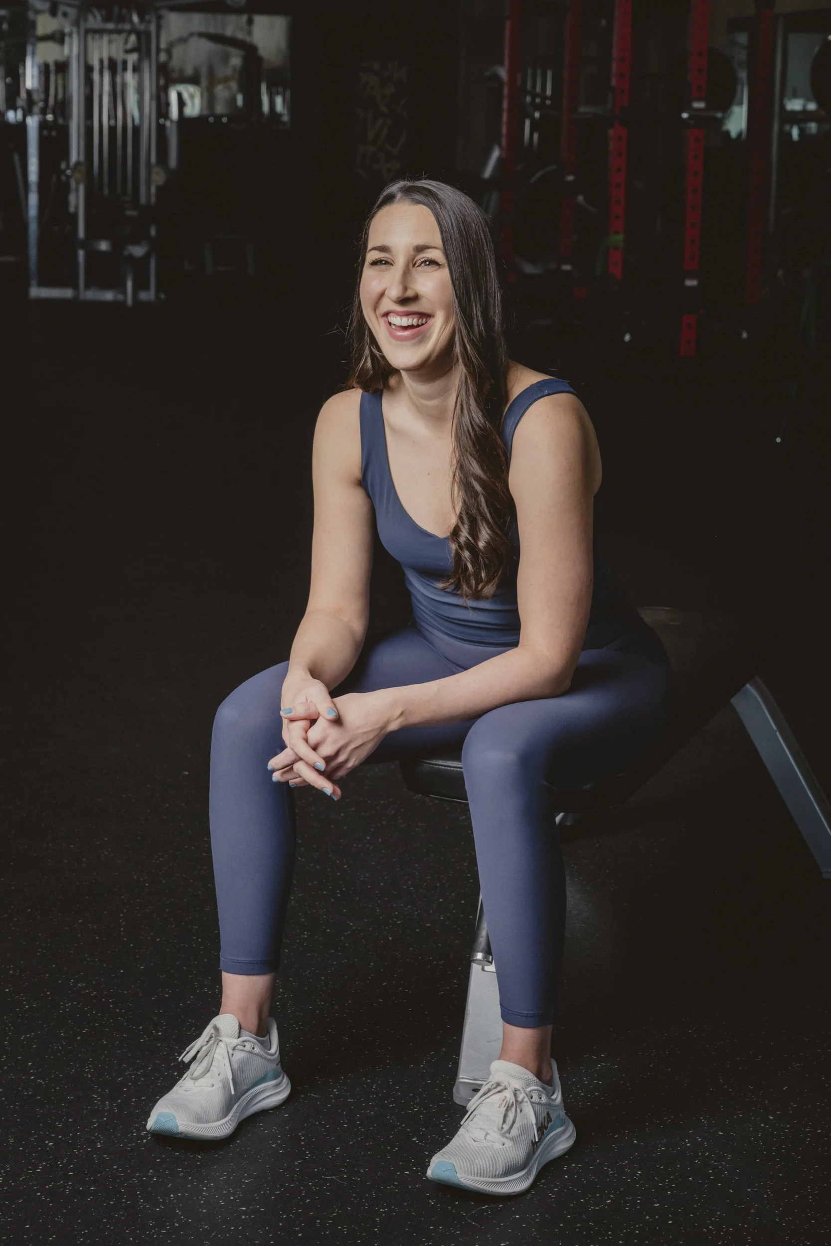 A woman sitting on a bench in a gym, wearing a navy athletic outfit and sneakers, smiling