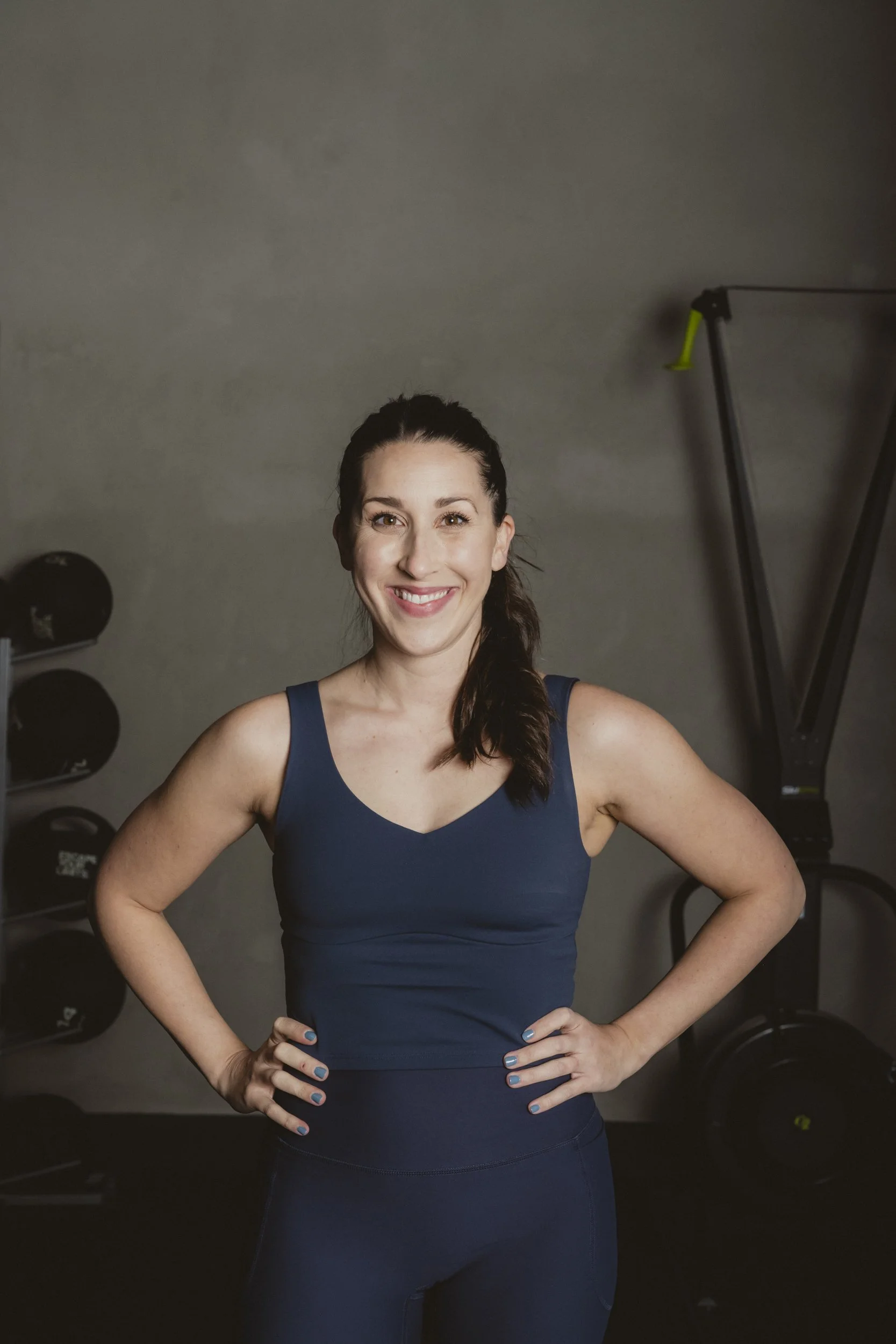 A woman in a navy workout outfit smiling with hands on hips in a gym, with weightlifting equipment in the background.