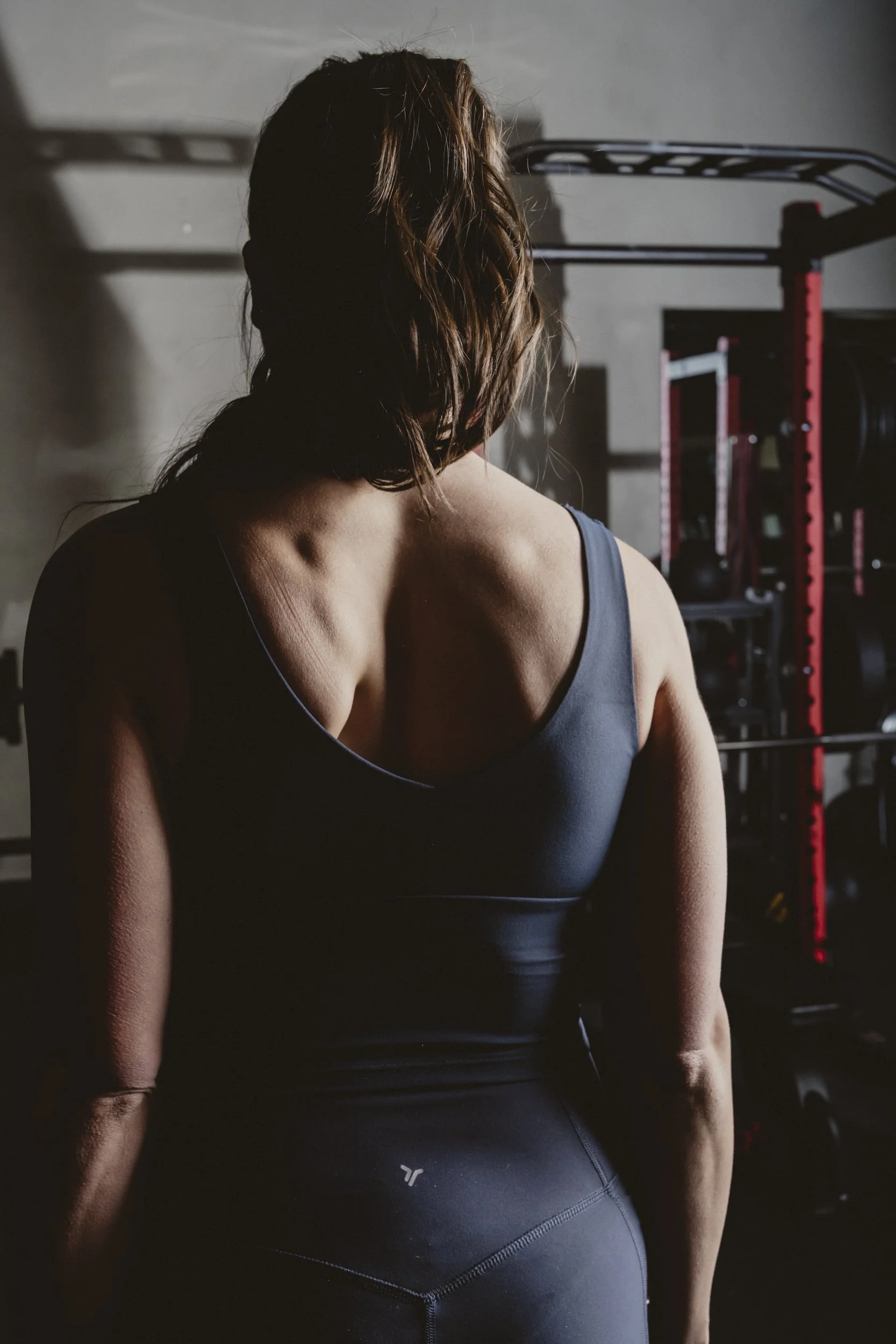 A woman with brown wavy hair in workout attire standing in a gym, facing away from the camera.
