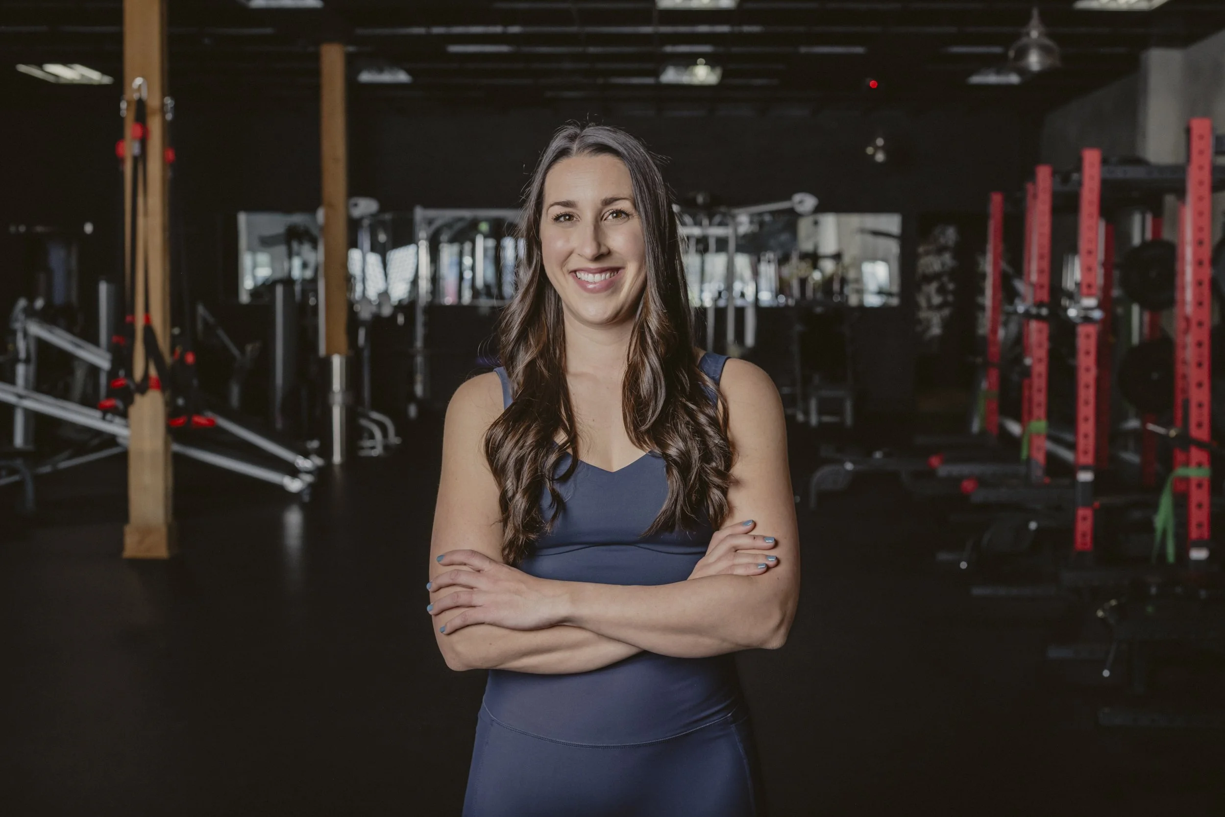 A smiling woman with long brown hair standing with her arms crossed inside a gym, with workout equipment in the background.