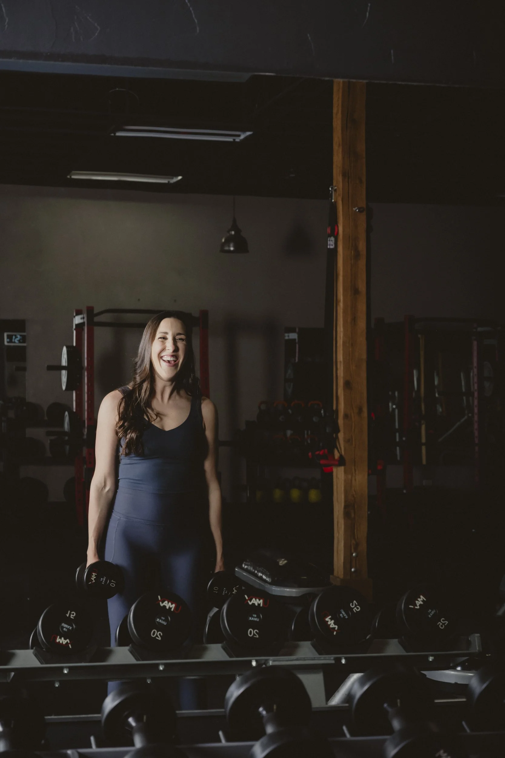 A woman in workout attire holding a dumbbell in a gym with various weights and fitness equipment in the background.