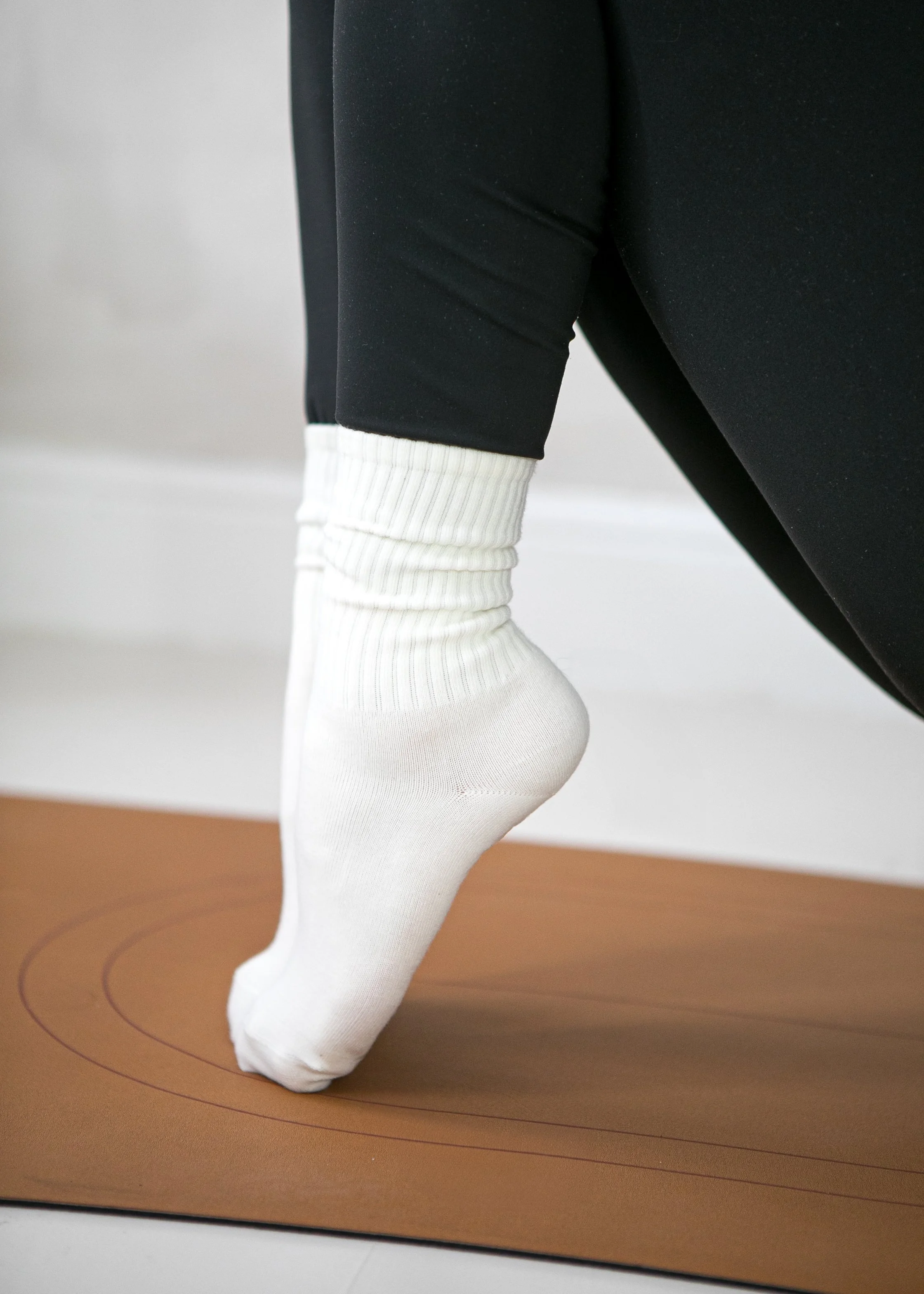 A person is doing pilates on a brown mat, with toes pointed on the mat, wearing black pants, white socks, and a black long-sleeve shirt.