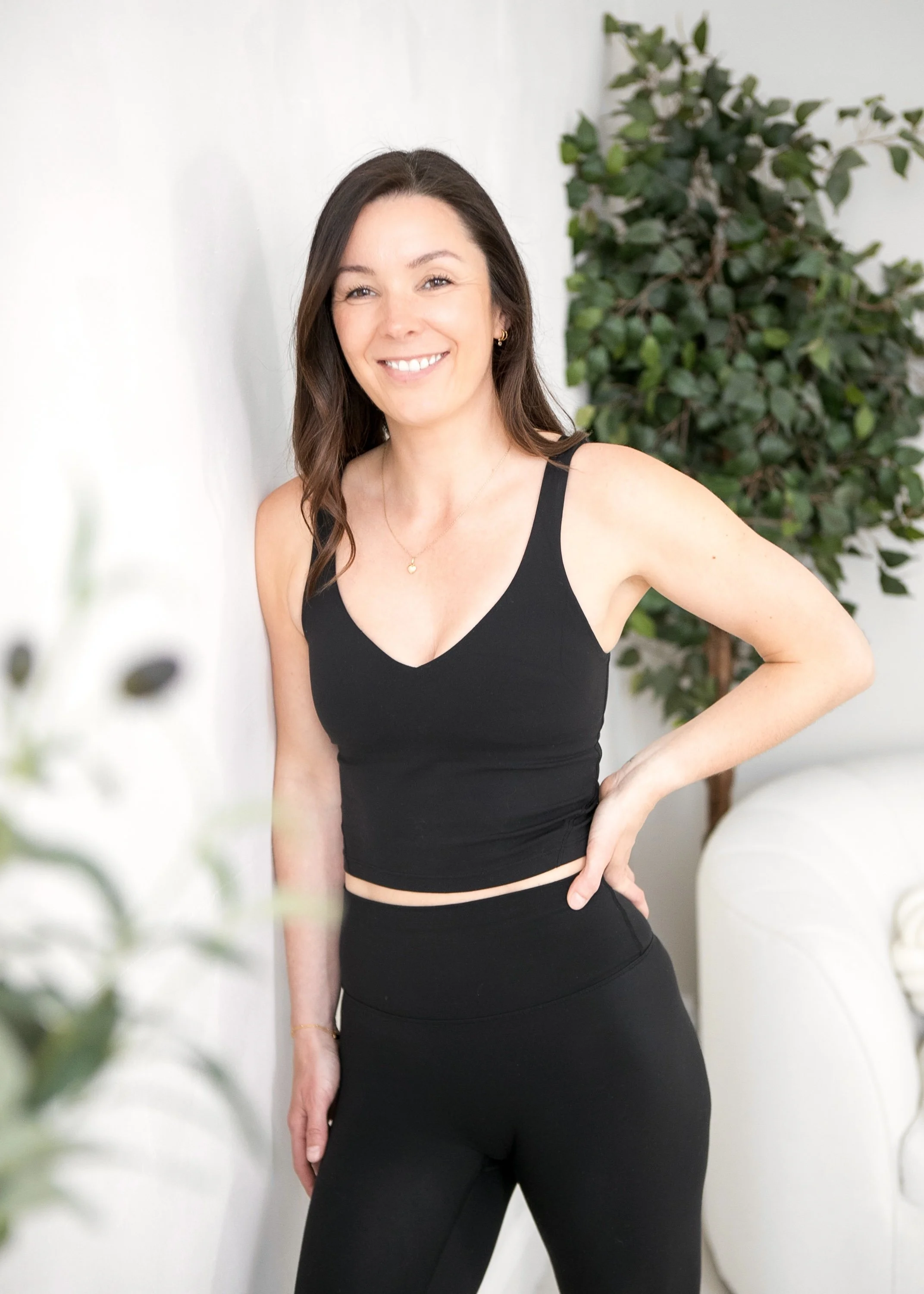 A woman with brown hair in a black tank top and matching black pants, smiling, standing indoors next to a white wall and a tall green leafy plant.