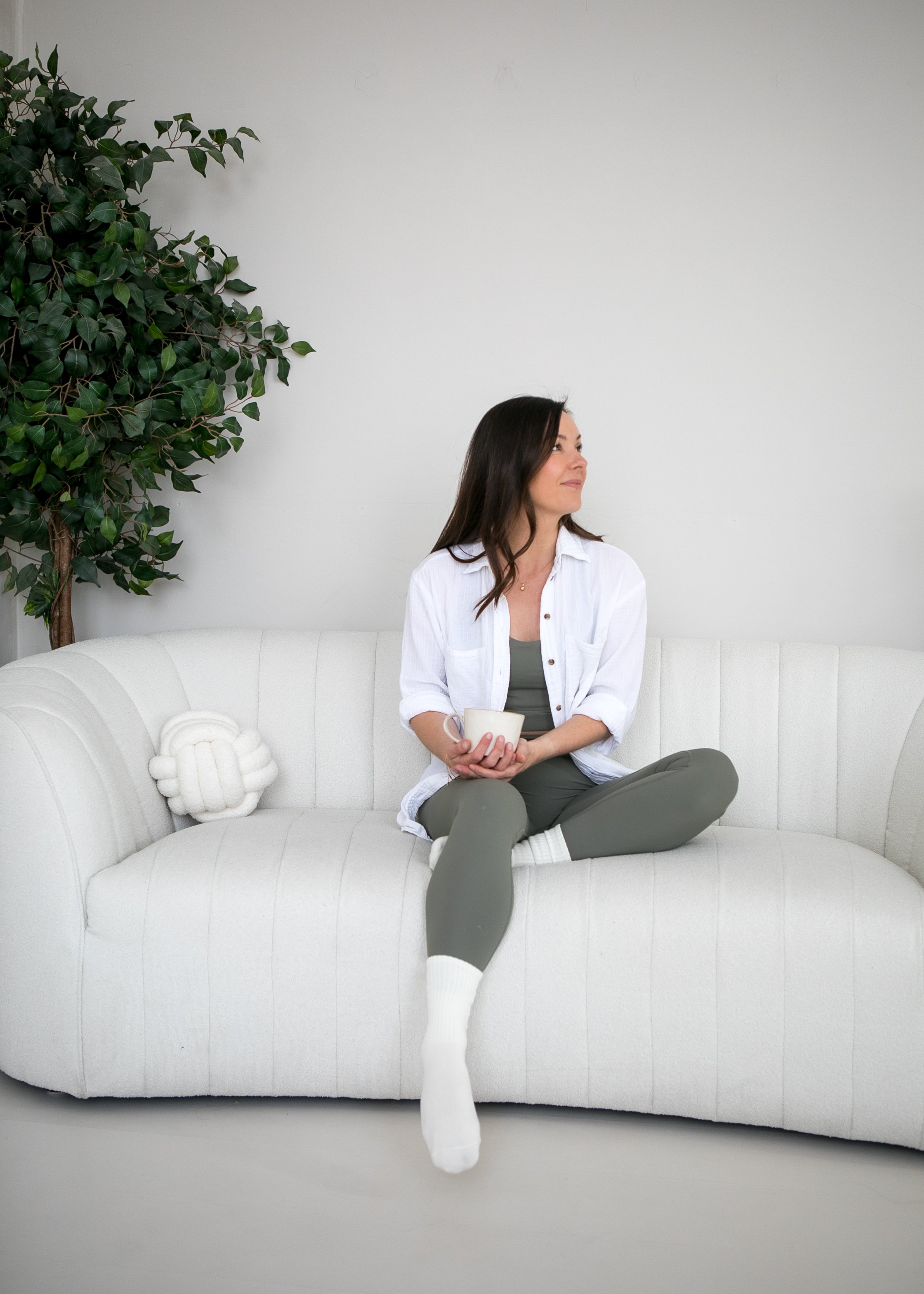 A woman sitting on a white sofa holding a mug, wearing a white shirt and sage green leggings, with a green plant to her left and a decorative knot pillow on the sofa.