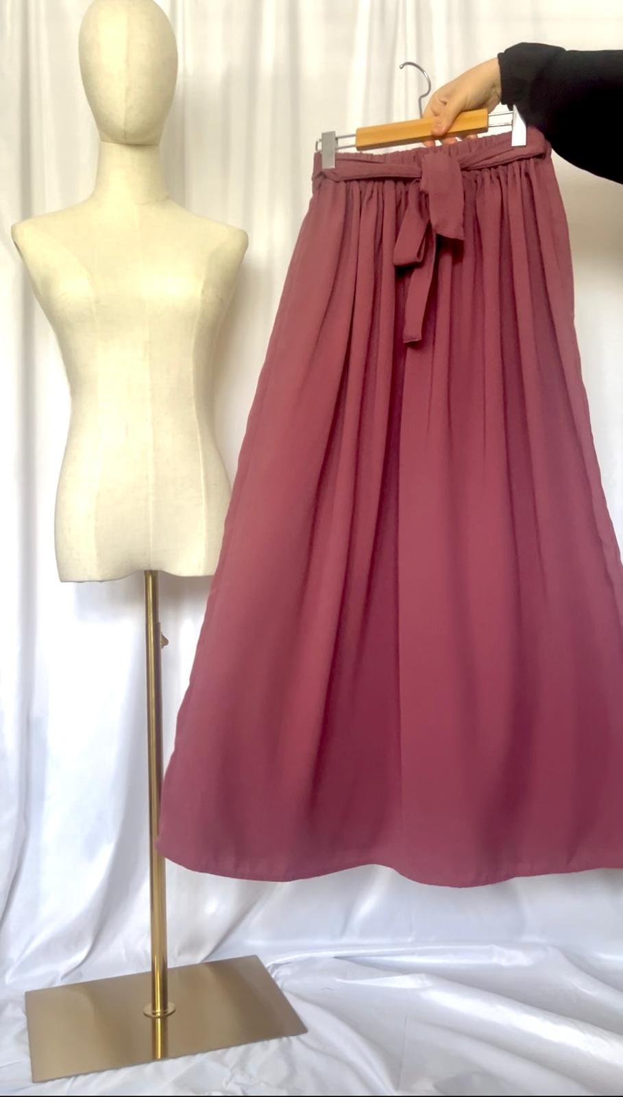 A mannequin is displayed next to a pink, high-waisted, pleated skirt with a bow tied at the waist.