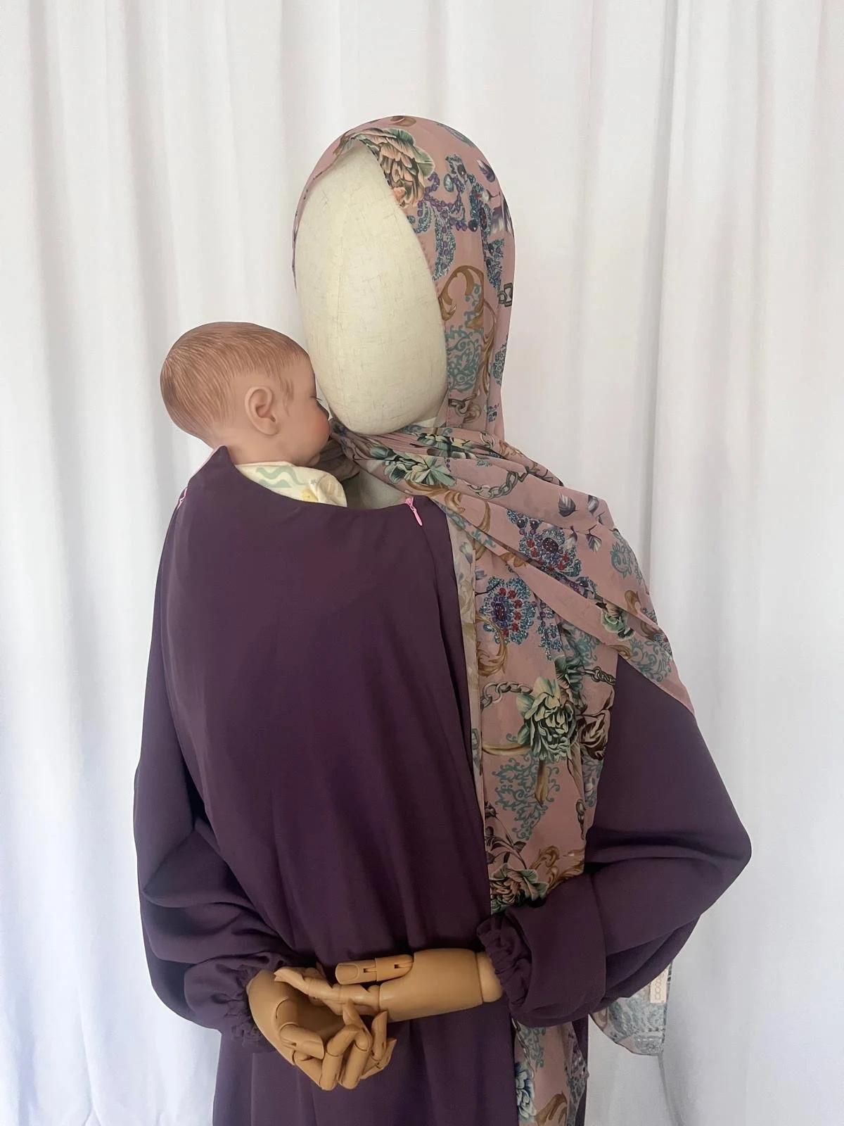 A mannequin dressed in a floral hijab and a dark lilac abaya, the mannequin is wearing a baby carrier in which there is a baby. The mannequin is displaying the baby wearing abaya.