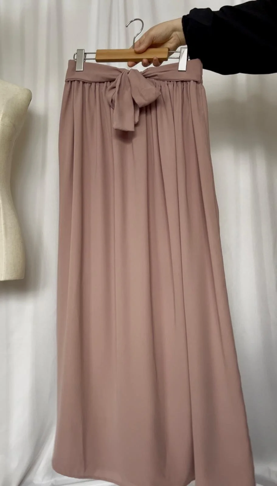 Long, dark cream pleated skirt with a bow at the waist hanging on a wooden hanger held by a person wearing a black sleeve.