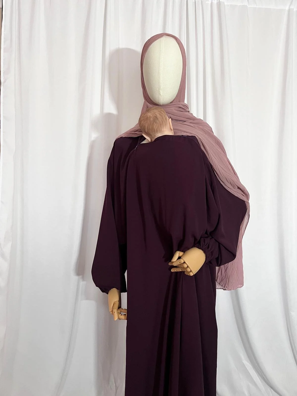 A mannequin dressed in a plum colored abaya with a light pink head covering, standing in front of white curtains. The mannequin is shown baby wearing with the baby wearing abaya.