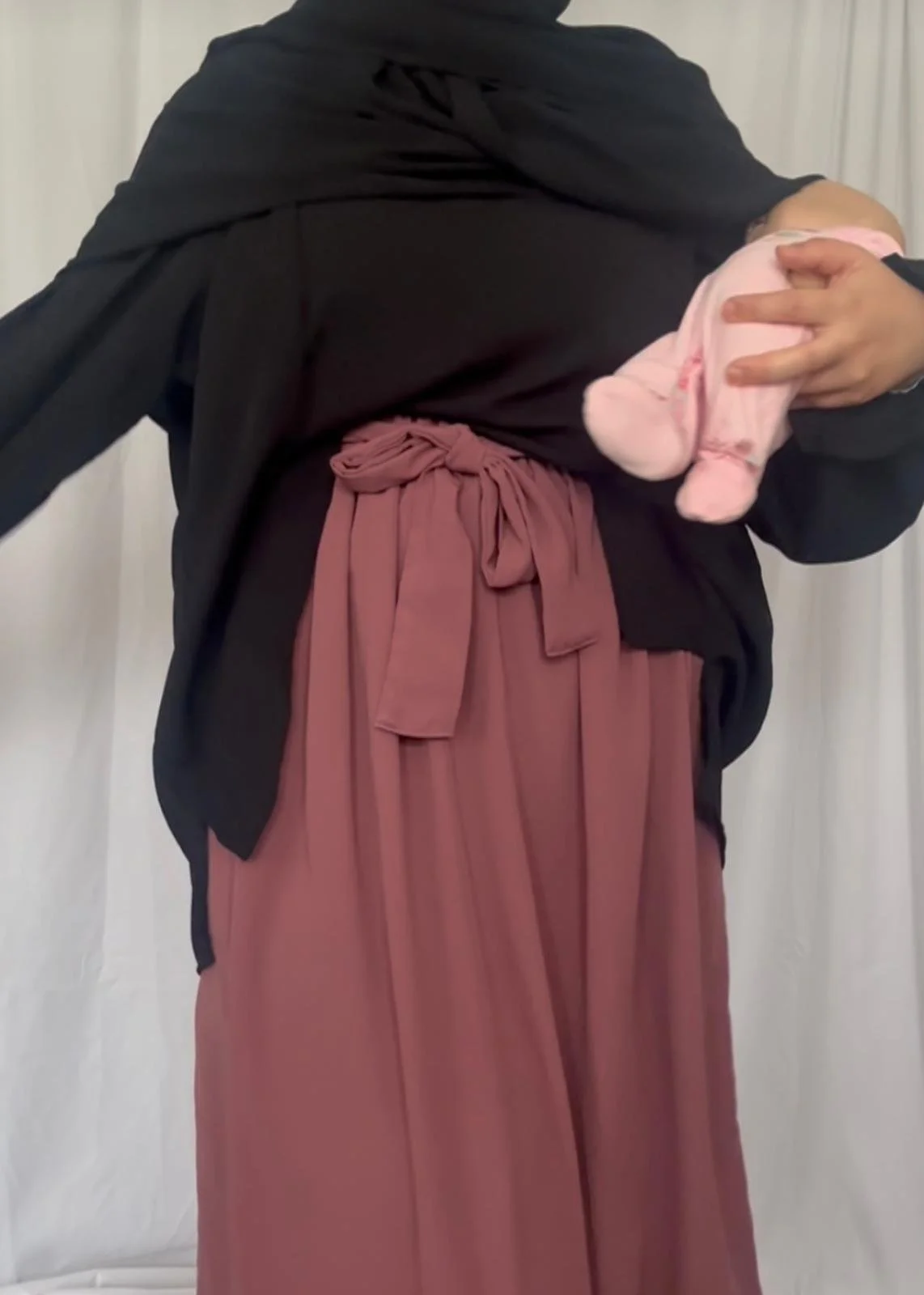 Person holding a baby in pink clothes, wearing a black top and rust-colored skirt with a tied waist.