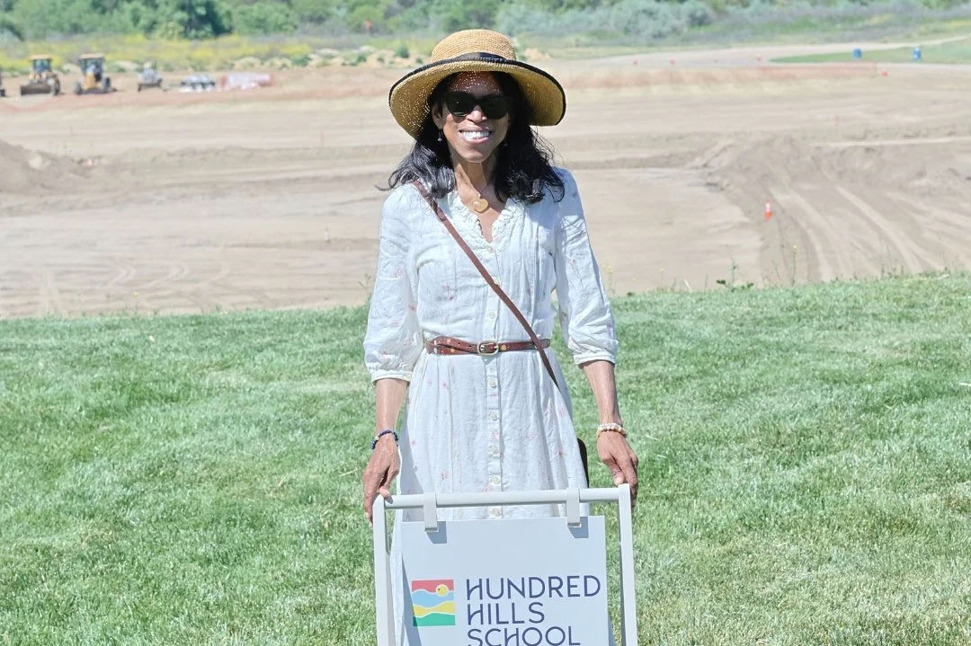 Woman wearing a wide-brimmed hat and sunglasses, standing outdoors on a grassy area, holding a sign that reads 'Hundred Hills School' on a sunny day with a construction or excavation site in the background.
