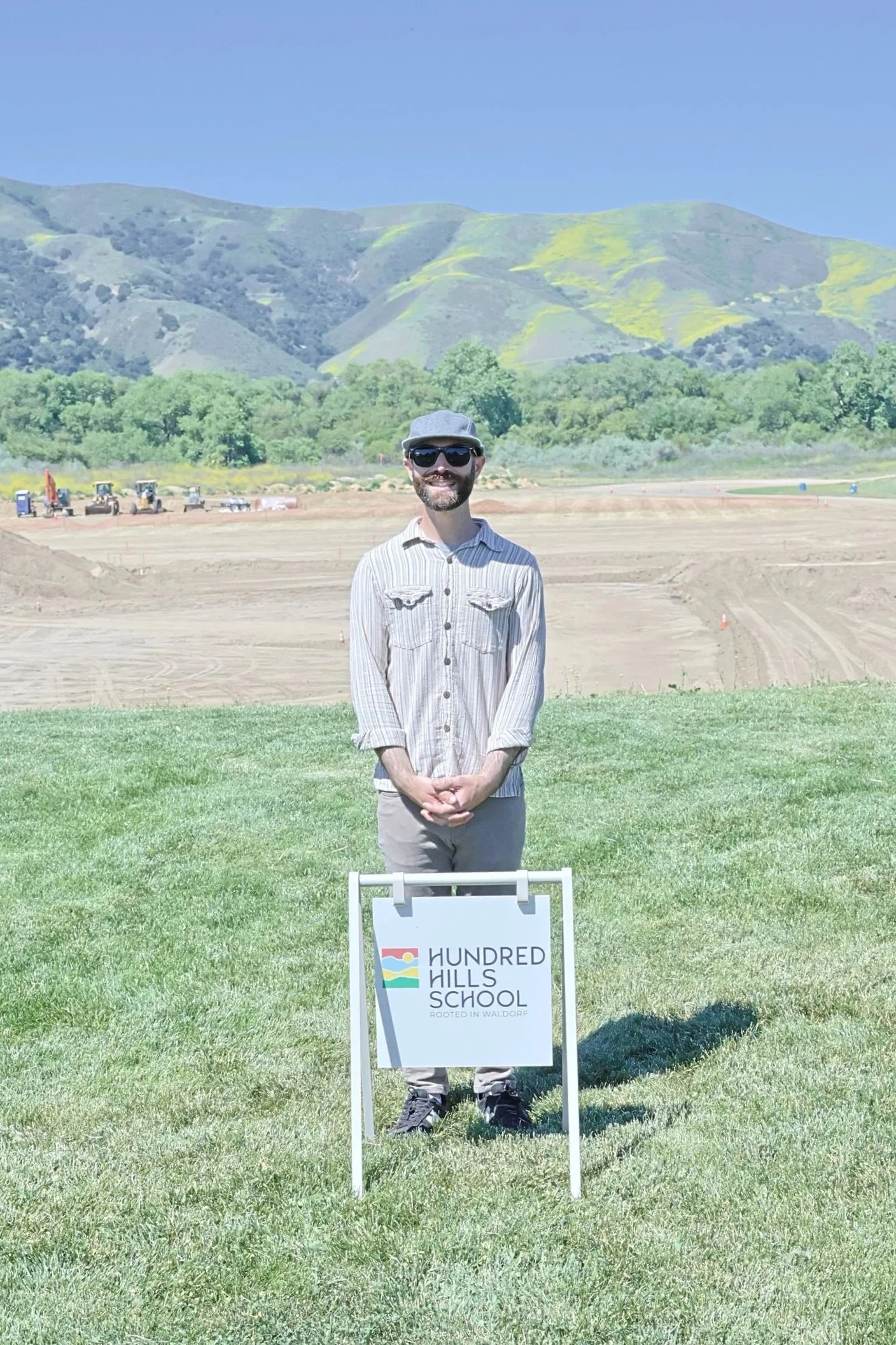 A man wearing sunglasses, a gray hat, and a striped shirt standing outdoors on a grassy area with mountains and construction equipment in the background. There is a sign in front of him that reads 'Hundred Hills School'.