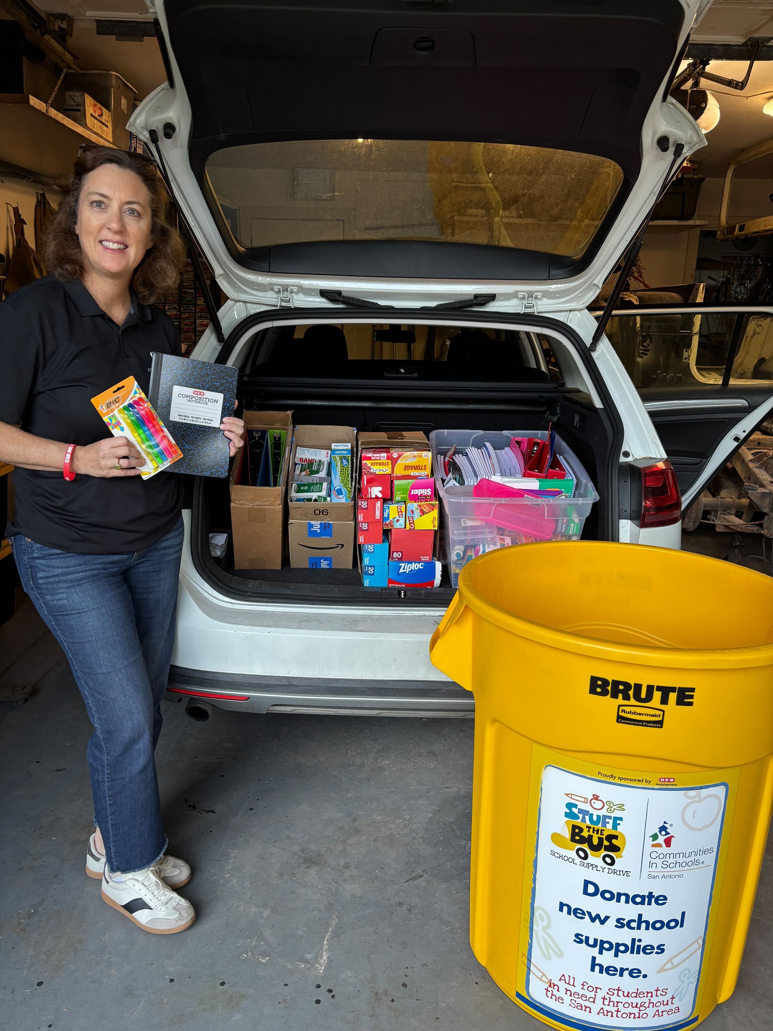 A woman standing beside an open car trunk filled with school supplies, holding a pack of markers and a composition notebook, with a yellow donation container labeled for school supply donations for a school drive.