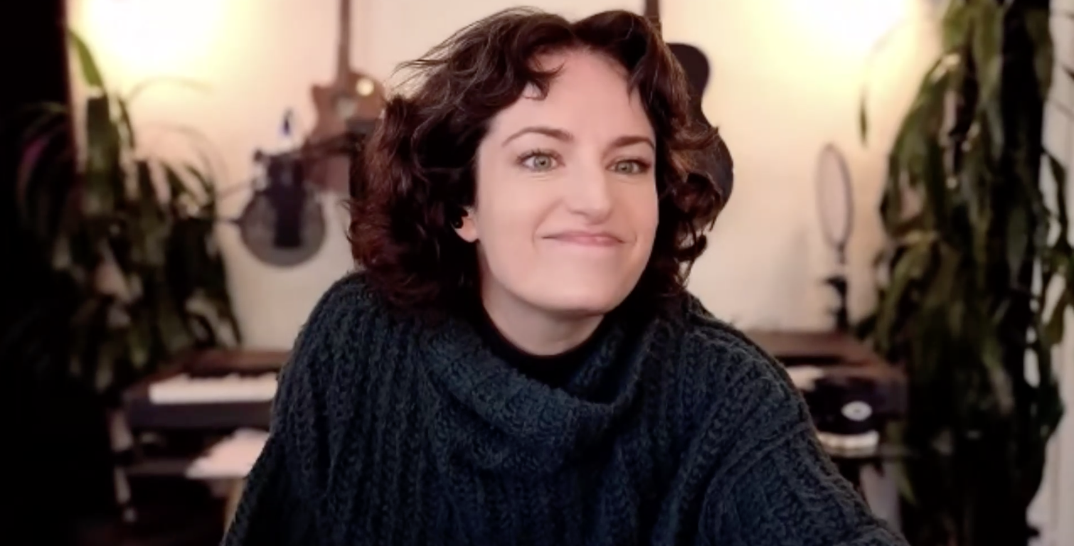 A woman with mid-length, curly brown hair, wearing a dark knitted sweater, smiling softly and sitting in a room with musical instruments and plants in the background.