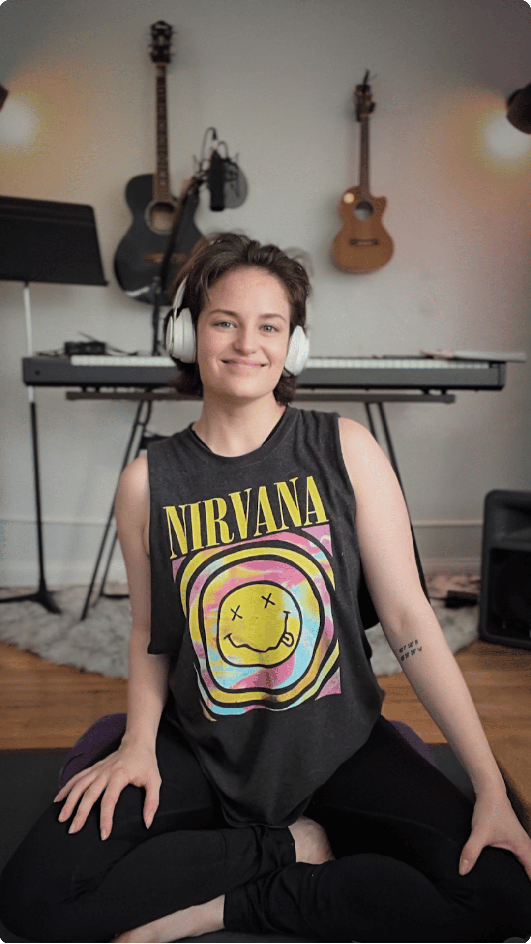 A woman with short hair smiling, wearing a Nirvana band tank top and headphones, sitting cross-legged in front of a music setup including guitars, keyboard, and music stand in a room with wooden floor and lights.