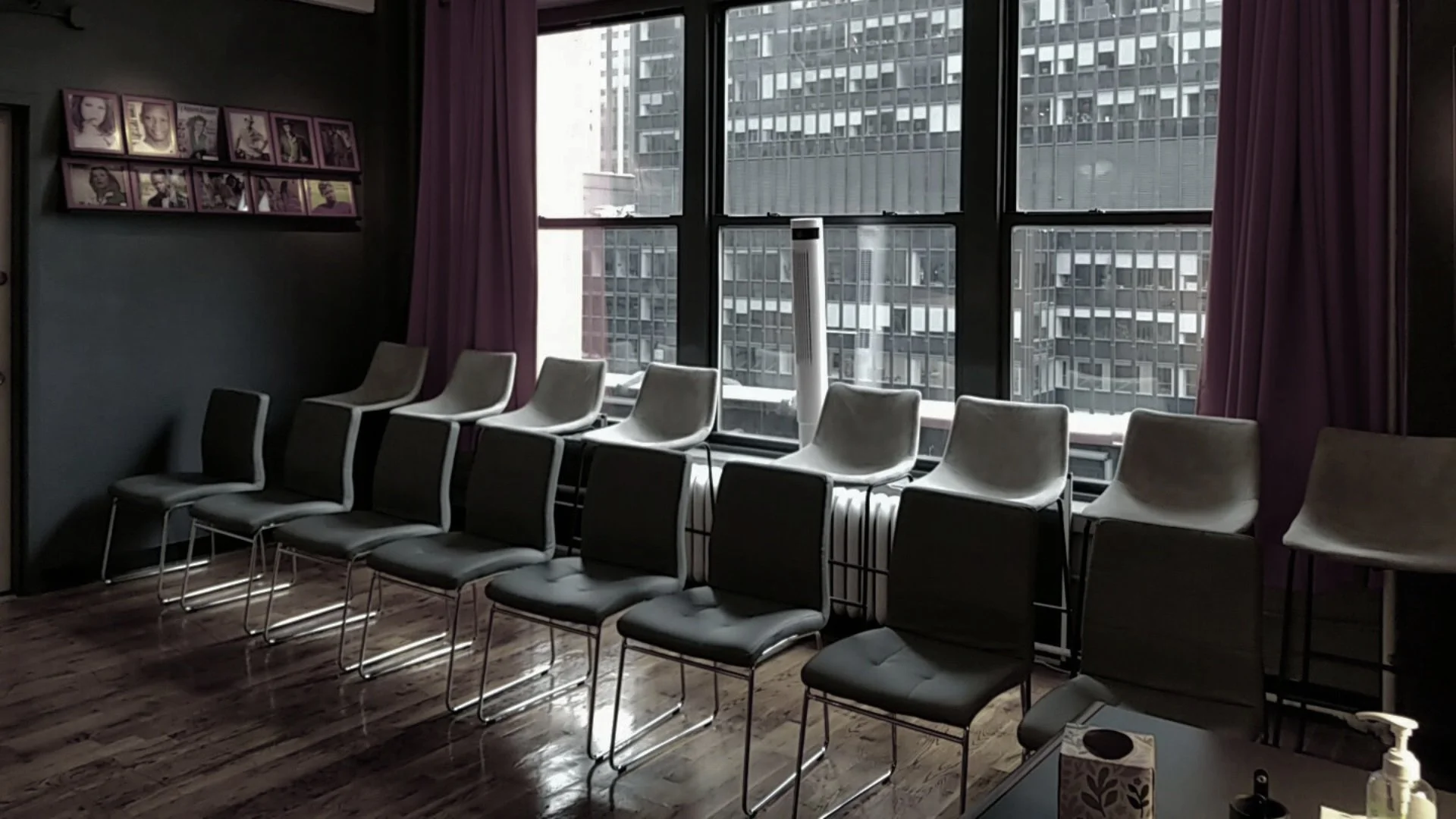 Empty room with stacked chairs near large windows showing a cityscape, purple curtains, and a photo collage on the wall.